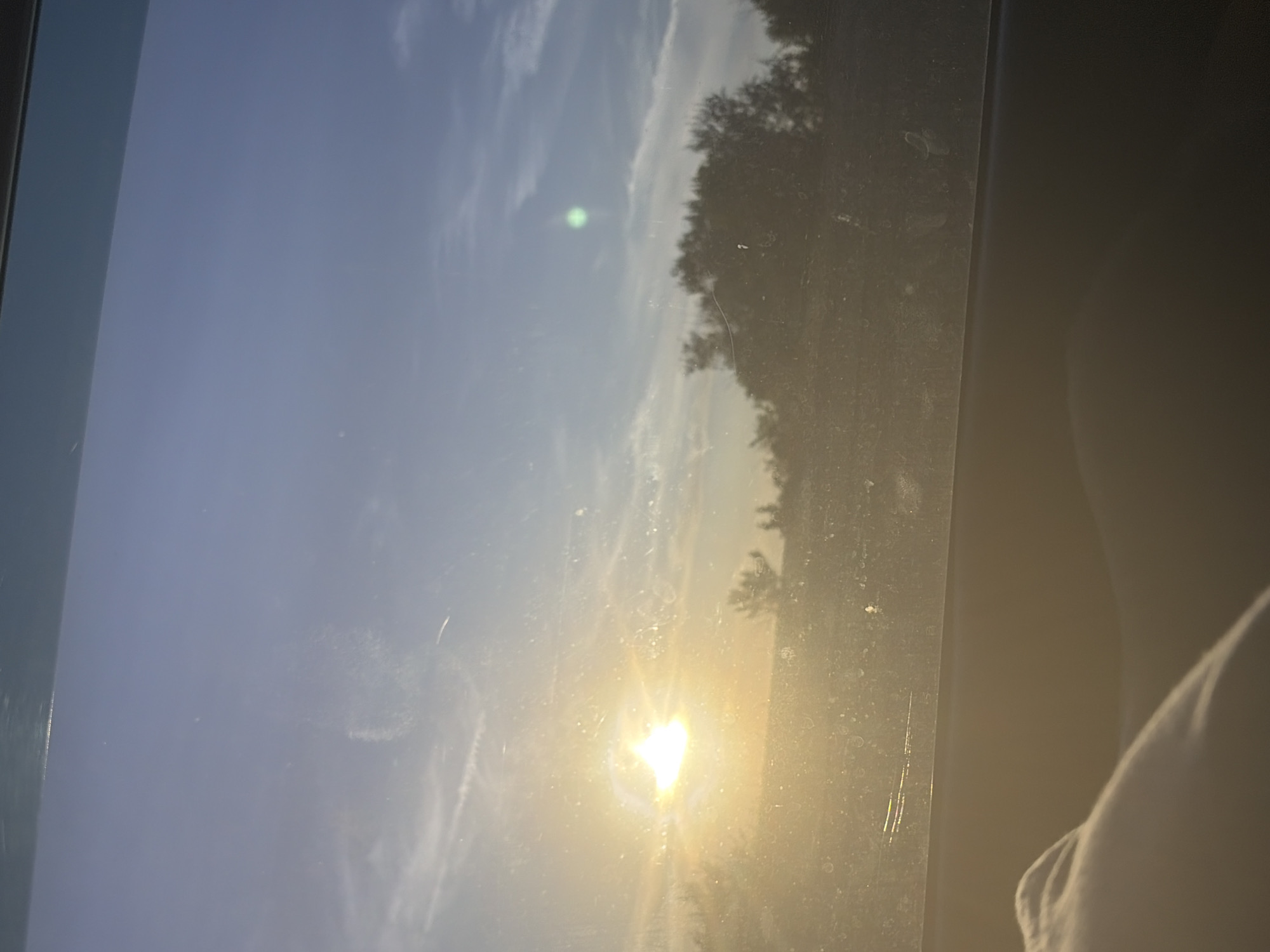 The image is a landscape captured through a window, likely from a vehicle, showing a sunset. The sun is a bright, luminous orb in the left half of the frame, casting a warm, golden glow over the scene. Wispy clouds streak across a blue sky, transitioning to a deeper shade towards the top. In the middle ground, silhouetted trees and vegetation create a dark, textured horizon. The foreground is dominated by a hazy, out-of-focus area, possibly the interior of the vehicle or an object obstructing the view. There is no discernible activity or interaction among people or subjects within the frame. The overall impression is one of a serene, natural environment at dusk. The location in Belogorki, Russia is not visually identifiable from the image itself.