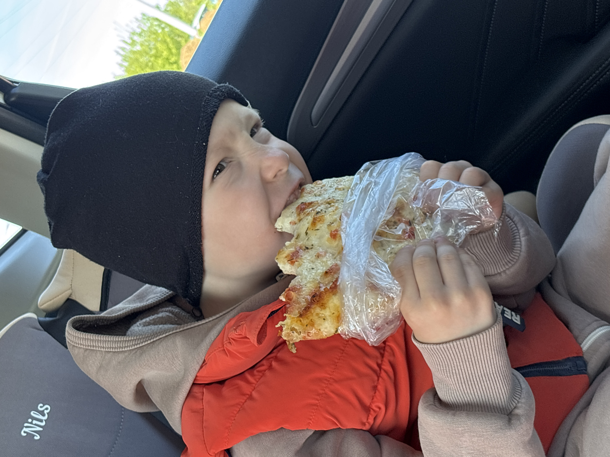 A young child, wearing a black knit cap, a light brown hoodie with a red vest underneath, is seated in a car seat. The child is eating a large slice of pizza that is wrapped in plastic. "Nils" is written on the side of the car seat. The location is unknown but the city listed is Chaikovsky, Russia.
