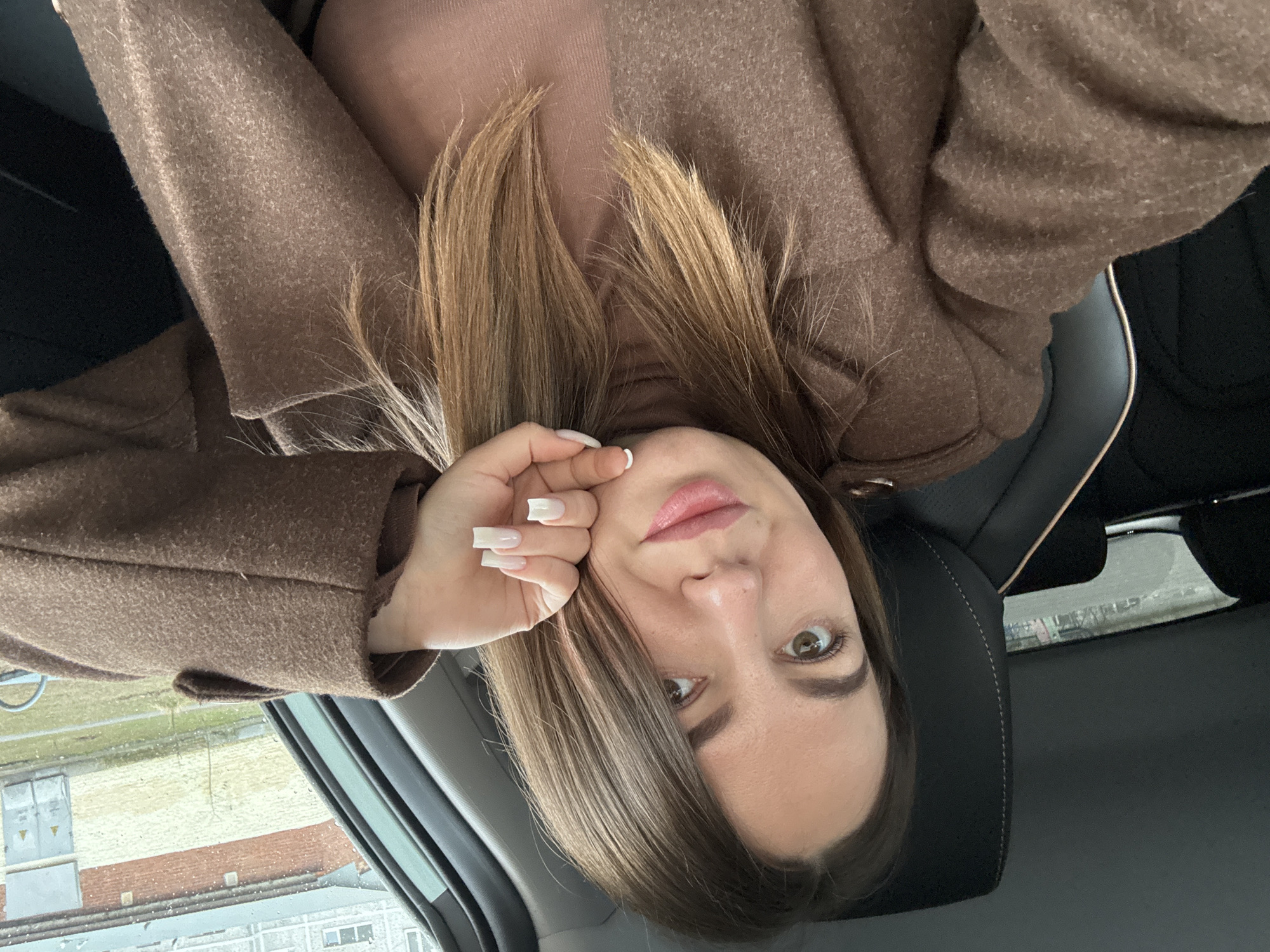 A woman is taking a selfie. She is wearing a brown coat over a light pink turtleneck. She has light brown hair with blonde highlights. She is holding her hand up to her face. The background shows a building and a gray sky. The woman is inside of a car, and it is raining. The location is likely Likino-Dulyovo, Russia.