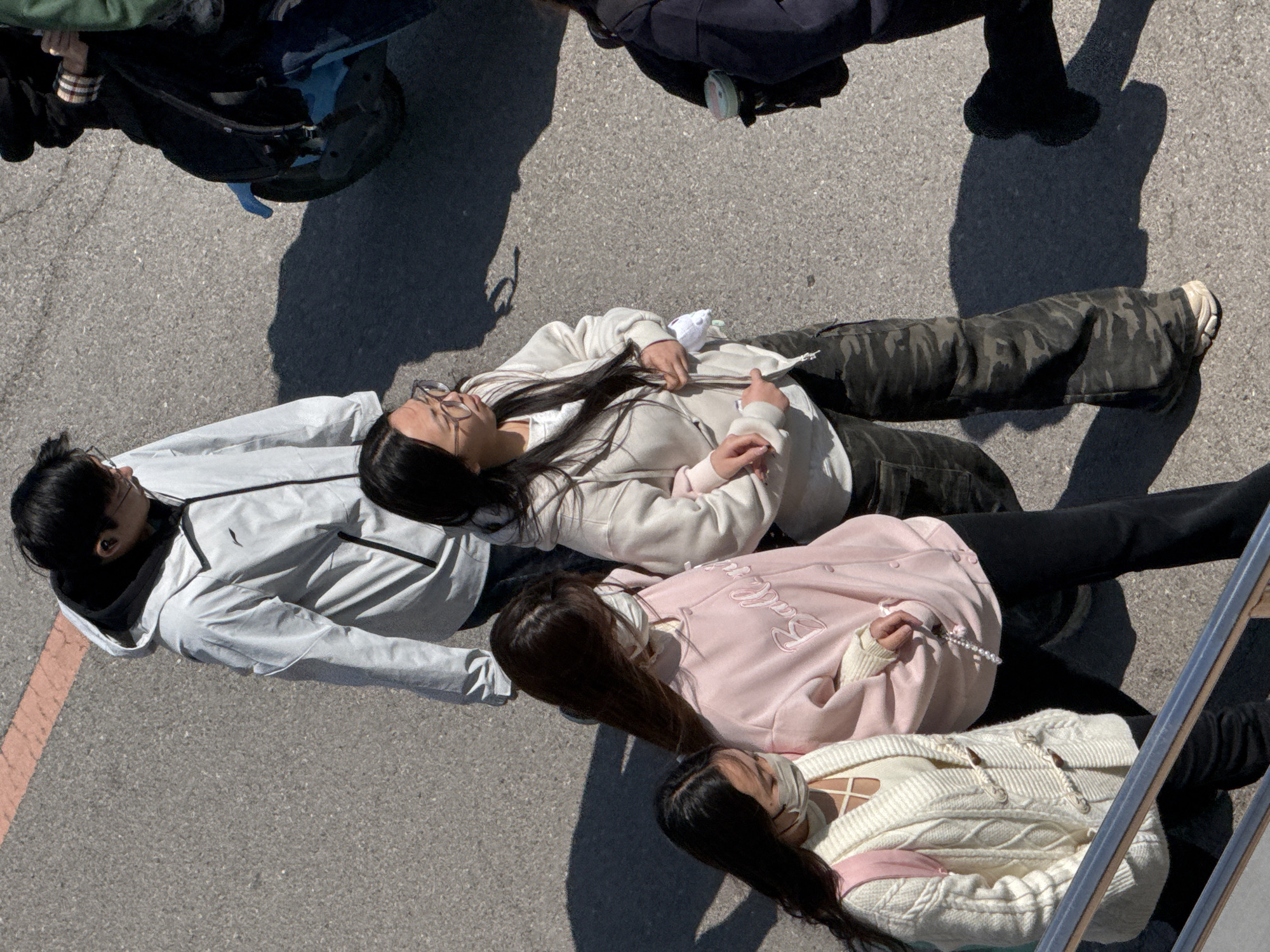 The photograph shows four young women lying on an asphalt surface, possibly a parking lot or street. The view is from an elevated position, looking down. The woman at the top is wearing a light beige jacket, camouflage pants, and glasses. The next woman is wearing a light pink jacket with writing on it and black leggings. The next woman is wearing a white cardigan. The first woman is wearing a white jacket and black leggings.
A red line is visible on the left side of the frame. The lighting is bright and sunny, casting shadows from people and objects outside the frame, suggesting the image was taken during the day. The photograph was taken in Zhongtun, China.