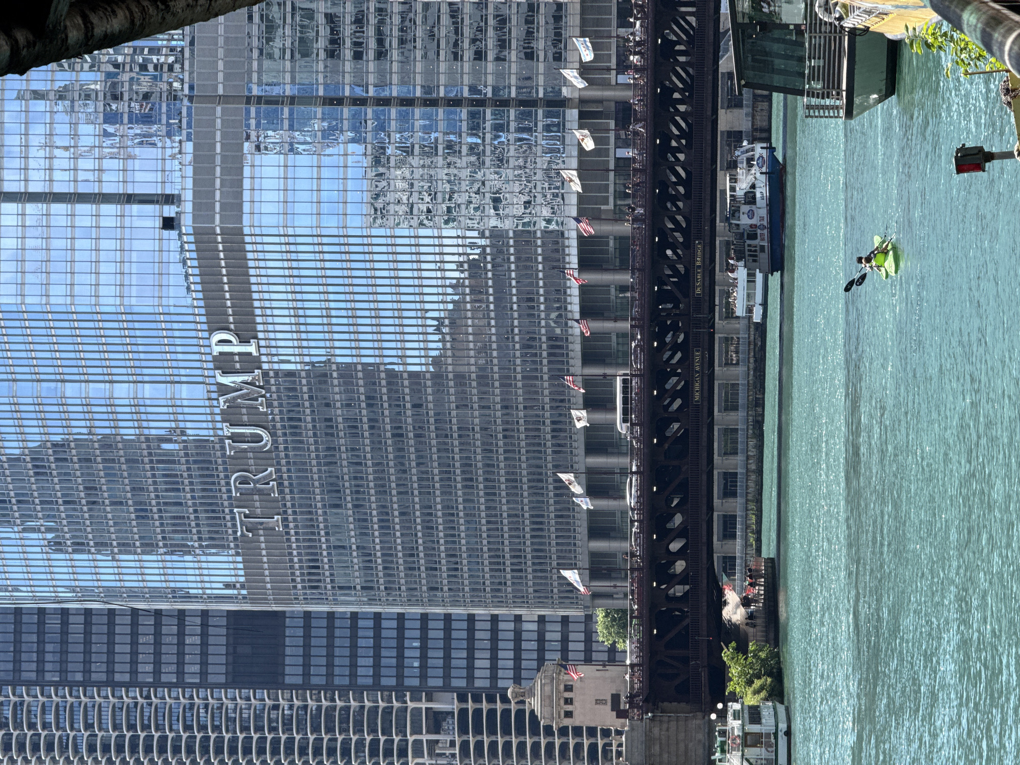 The image captures a view of the Trump International Hotel and Tower in Chicago, with the Chicago River flowing alongside. The towering skyscraper, characterized by its glass facade reflecting the sky, dominates the left side of the frame. The word "TRUMP" is prominently displayed in large, metallic letters on the building's exterior.
On the river, a person is kayaking in a bright green kayak, paddling through the shimmering turquoise water. A tour boat is also visible on the river, with people on its deck. A bridge, part of the city's infrastructure, spans the river and is adorned with several flags, including American flags, fluttering in the breeze. The architecture of the bridge suggests a historical element within the modern urban landscape.
The scene is illuminated by natural light, indicating it is daytime, likely a sunny day given the bright reflections and the clear water. The overall atmosphere is one of urban activity and recreation. Text visible on the bridge reads "DUSTABLE BRIDGE" and "MICHIGAN AVENUE," providing specific location cues. The presence of people engaged in different activities—kayaking, being on a tour boat, and observing from the bridge—suggests a vibrant and dynamic environment in the heart of Chicago.