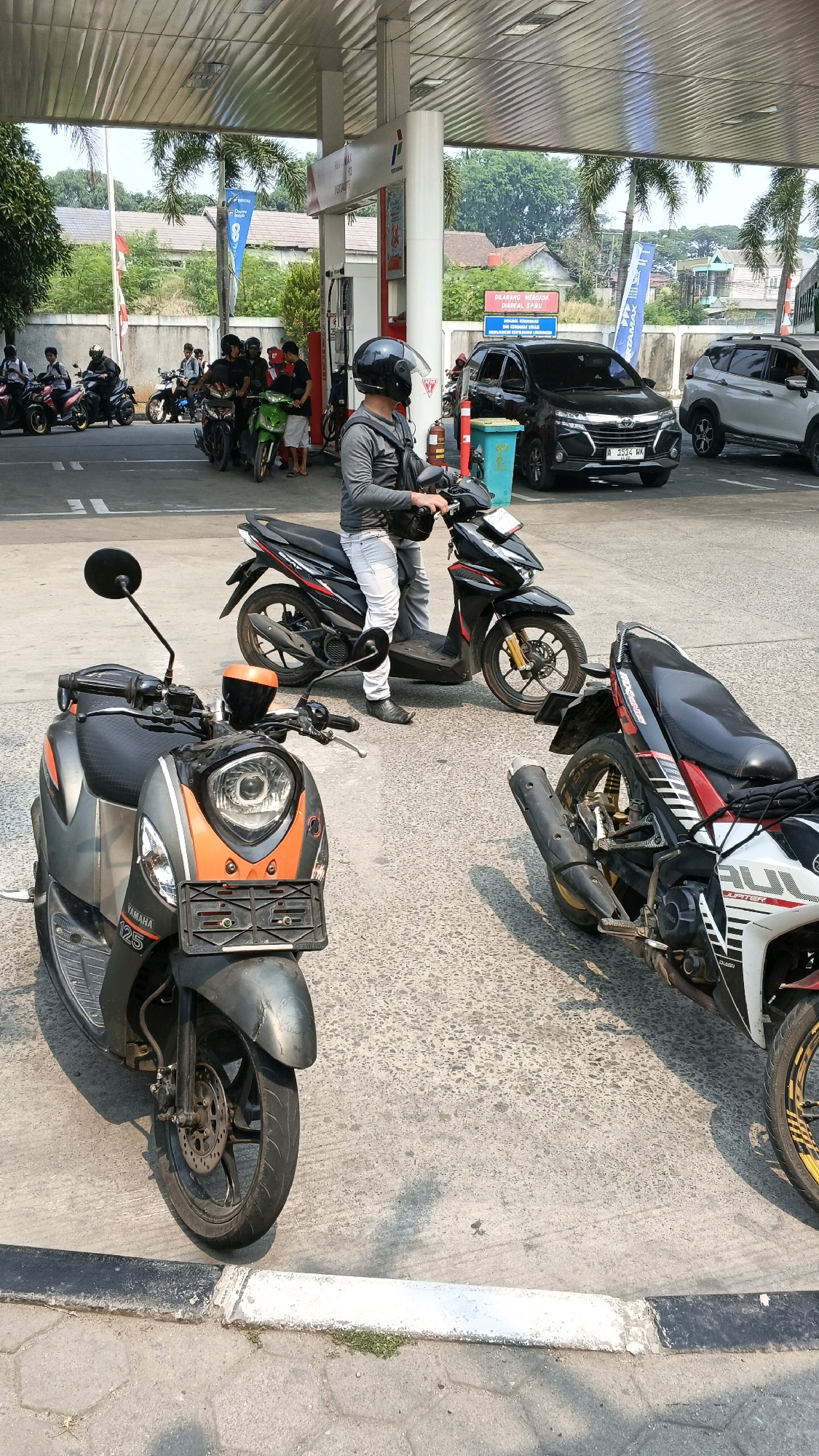 This is a medium-shot, eye-level, outdoor photograph taken at a Pertamina gas station in Balaraja, Indonesia. The foreground features three parked motorcycles. The closest motorcycle, on the left, is a silver and orange scooter with "YAMAHA 125" visible on its side. To the right of this scooter is a black and red motorcycle. Further back and to the right is another motorcycle.
In the middle ground, a person wearing a gray long-sleeved shirt, white pants, black shoes, and a black helmet is standing next to a black scooter, holding onto its handlebars. Behind this person, several other people on motorcycles are visible, appearing to be waiting or refueling. A black SUV with license plate "A 1514 HK" is parked behind the central figures. To the right of the SUV, a white SUV is also visible.
The background shows a gas station canopy with white poles and a reflective ceiling. Palm trees and buildings are visible behind the canopy. A blue banner with white text is hanging from a pole on the left side of the image. The ground is paved with asphalt and concrete. The lighting suggests it is daytime, with shadows cast on the ground.