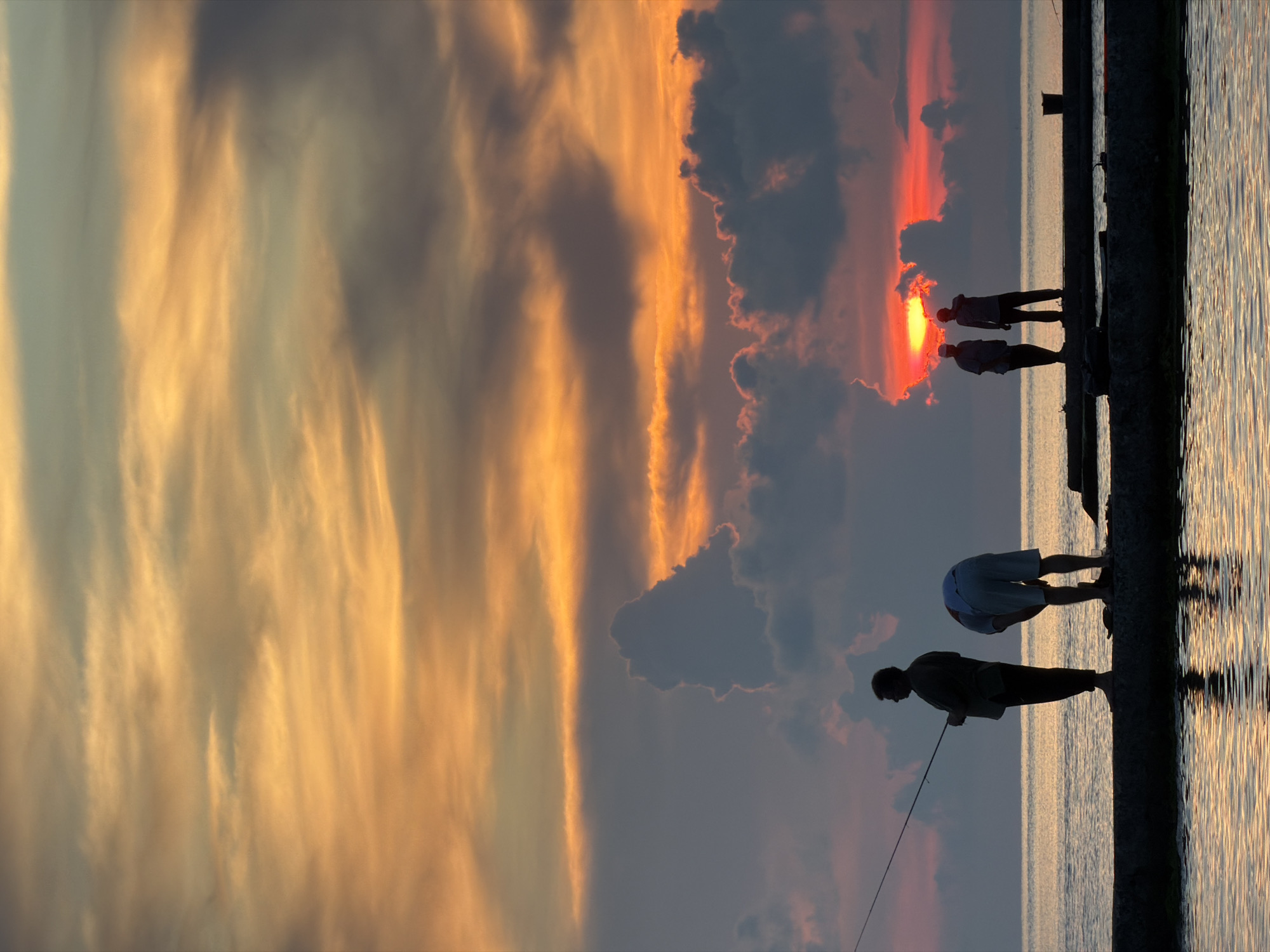 The image depicts a sunset scene at a coastal location, likely the city of Dederkoy, Russia. Silhouetted figures stand on a concrete pier extending into the water. On the left, a person is actively fishing with a rod. Near the center, another individual is bent over, possibly attending to something on the pier. Further to the right, two people are conversing. The sky is ablaze with warm hues of orange and yellow, characteristic of a sunset, with clouds tinged with red. The water reflects the vibrant colors of the sky. A bucket is visible on the pier in the background. The overall atmosphere is serene, capturing a moment of leisure at dusk.