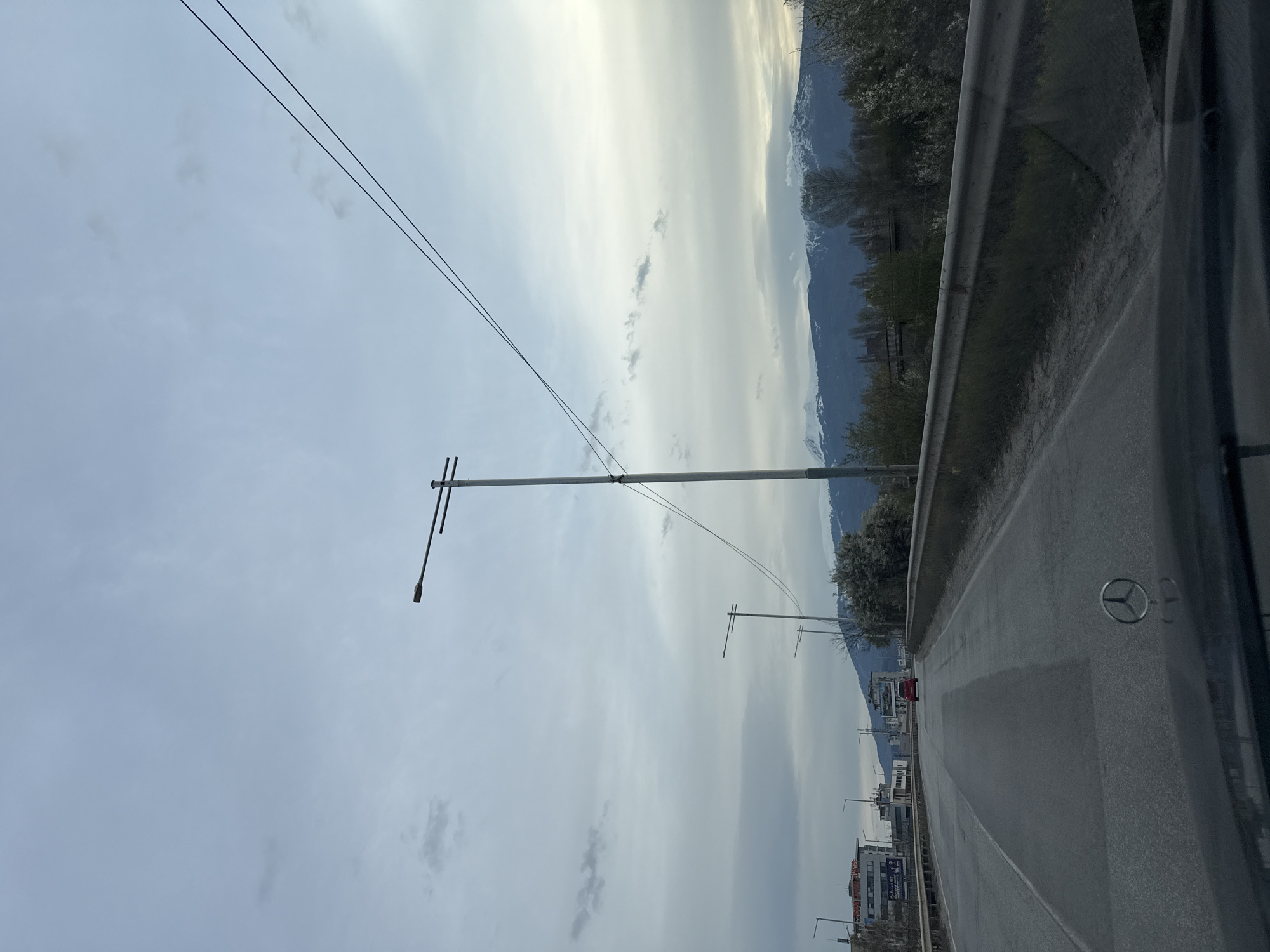 The image depicts a view from inside a vehicle, likely a car, traveling along a road on the outskirts of Sofia, Bulgaria. The road is paved and features a median line. A metal guardrail runs along the right side of the road. The vehicle's window and the Mercedes-Benz logo are visible on the right side of the frame, suggesting the car's make.
Beyond the road, a line of trees and vegetation separates the road from a mountainous region. A series of streetlights line the left side of the road, along with some buildings. Overhead power lines stretch across the sky. The sky itself is overcast, with varying shades of gray and white clouds. A red vehicle is further down the road.