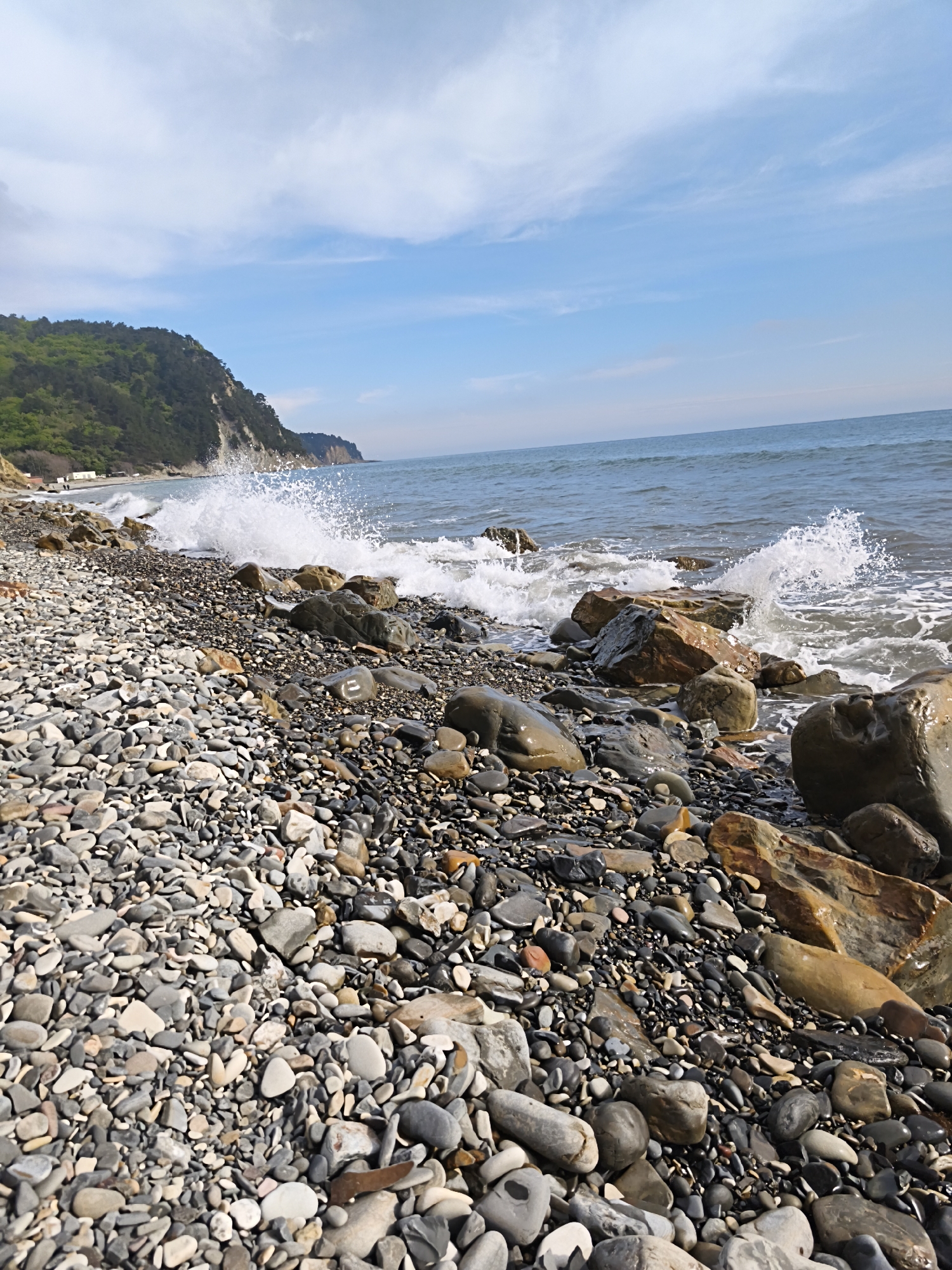 The image depicts a rocky beach in Praskoveyevka, Russia. The foreground is dominated by an expanse of gray and brown pebbles. Larger rocks are scattered along the shoreline, with waves crashing against them, creating white spray. In the background, a green hill rises, bordering the beach. The sky is light blue with scattered white clouds. The sea stretches to the horizon.