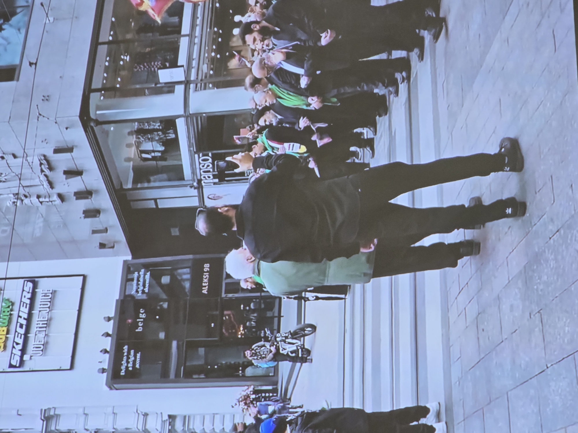 The image depicts a street scene in Helsinki, Finland, likely on Aleksanterinkatu given the sign for "ALEKSI 9B" visible on a building. In the foreground, a group of men are gathered. One man in the center is wearing a dark long-sleeved shirt and dark pants, with a green scarf draped around his shoulders. He appears to be holding a phone or camera up, possibly taking a photo or video. Behind him, other individuals are also present, some wearing dark clothing and head coverings.
Further back, on the left side of the frame, a man is seated on a white electric motorcycle, wearing a grey jacket and a backpack. Above him, prominent signage indicates "SUBWAY", "SKECHERS", and "JOE & THE JUICE". The buildings in the background feature large glass windows, suggesting commercial establishments. One window displays clothing on mannequins. The street is paved with stone tiles. The overall atmosphere appears to be that of a public gathering or event in an urban environment.
