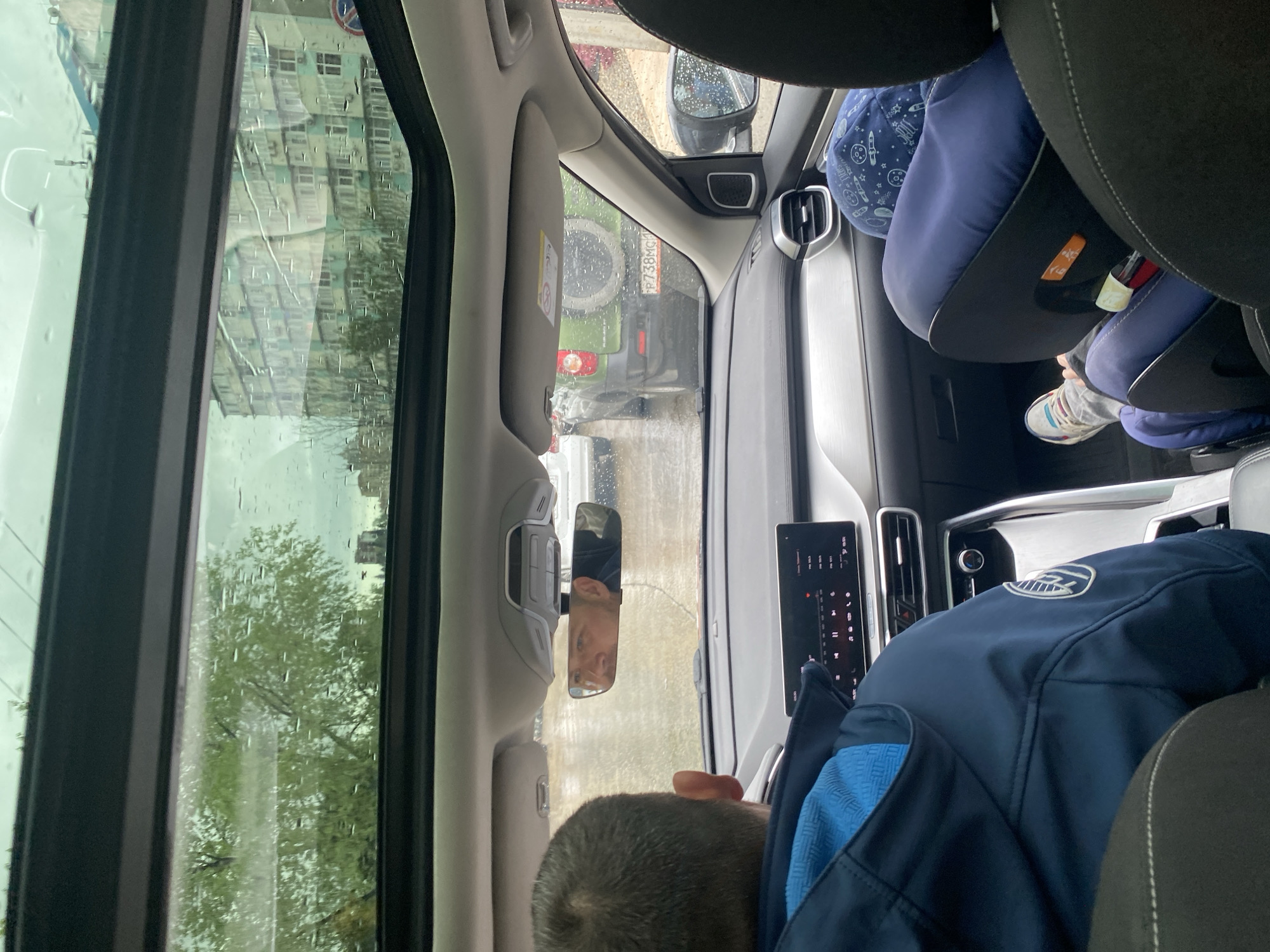 Here is a description of the image:
The image is taken from inside a car, looking forward through the windshield and sunroof, on a rainy day in Sochi, Russia. A child in a car seat with a blue patterned hat is visible in the back. A person wearing a dark blue jacket sits in the front passenger seat. The rearview mirror shows the reflection of the driver, and through the windshield, another green vehicle with the license plate "P738MG" is visible. The buildings outside are indistinct due to the rain. The dashboard of the car and various interior features are also visible.