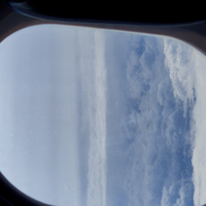 The image presents an aerial view of stratified cloud formations, consistent with an aircraft in flight over the airspace of Blagnac, France, or its vicinity. The scene is framed by the dark, curvilinear edges of an aircraft window visible on the left and right. The external view comprises three primary atmospheric layers: a lower, dense stratum of lumpy cumulus clouds in white and light grey; a distinct, flatter, uniform horizontal band of grey cloud positioned centrally; and an uppermost section of lighter, hazy blue-grey sky or very thin cloud cover. No ground features, individuals, or other specific objects are visible within the frame, focusing solely on the atmospheric environment.