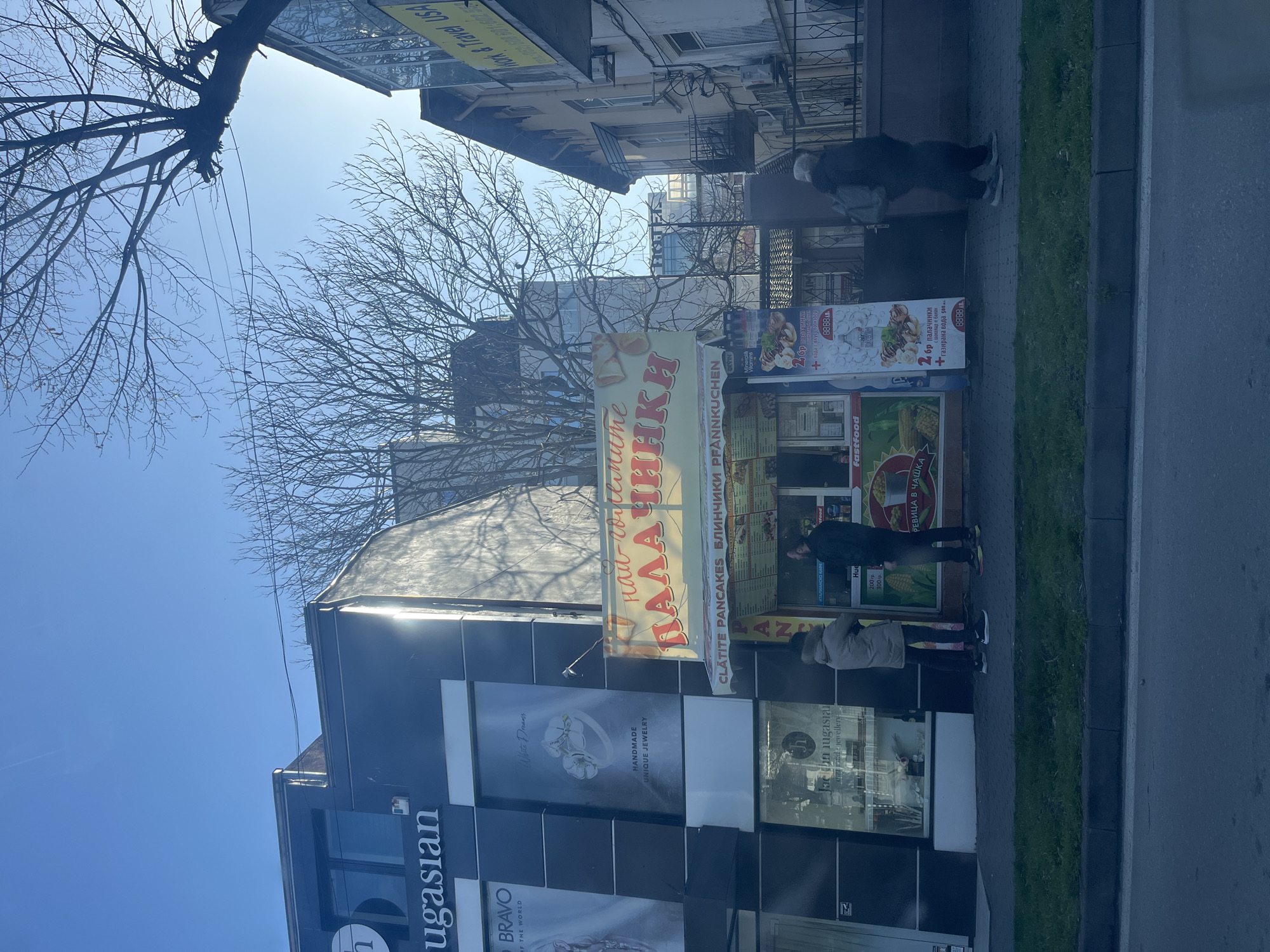 The image depicts a daytime urban street scene in Varna, Bulgaria, viewed from slightly below street level.
On the left, a modern, dark-paneled building houses "Jacsan Lugasian Jewelers," identifiable by "ugasian" signage and window displays featuring "HANDMADE UNIQUE JEWELRY" and "White Dreams." To its right, a brightly colored kiosk with a yellow awning prominently displays "най-вкусните ПАЛАЧИНКИ" (the most delicious pancakes) in Cyrillic, along with "CLATITE PANCAKES БЛИНЧИКИ PFANNKUCHEN" and various fast-food menu items.
Three individuals are visible on the sidewalk. Two individuals stand in front of the pancake kiosk; one wears a light-colored hooded jacket and faces right, while the other wears a dark jacket and faces left. A third person, dressed in dark clothing with light headwear, walks to the right, away from the kiosk, carrying a dark bag over their shoulder.
Further to the right, an older, light-colored building features multiple windows and a visible balcony. A partial sign on this building reads "Work & Travel USA." The street in the foreground consists of an asphalt road, a grey curb, a narrow strip of green grass, and a grey paved sidewalk. The upper portion of the image shows a clear blue sky, with the bare branches of several deciduous trees silhouetted against it. Bright sunlight is evident, casting reflections on the buildings.
