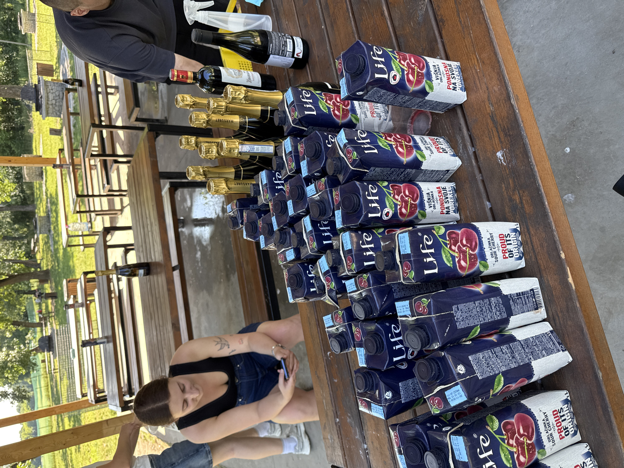 The image shows a wooden table with several bottles of wine and champagne lined up next to stacks of cherry juice boxes (brand “Life”), under a covered outdoor pavilion. A woman is sitting on a bench at the edge of the table, using her phone. Another person is standing at the edge of the table near the beverages, and a man wearing a black shirt stands at the other end. In the background, there are more tables with benches, and a grassy area with trees. It's likely in the city of Cukarica Urban Municipality, Serbia, based on the label of the cherry juice box.
