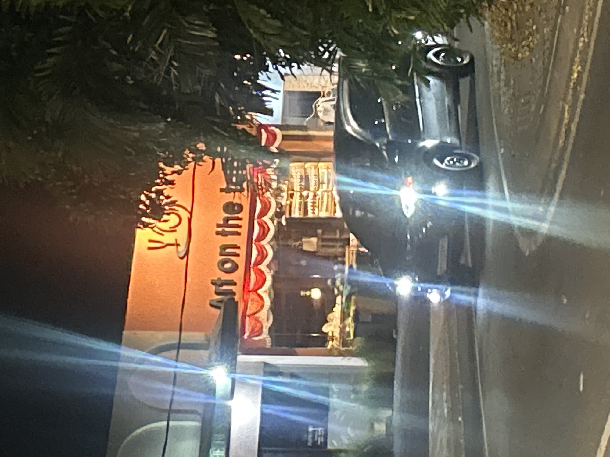 The image depicts a street scene at night in North Kuta, Indonesia. A dark-colored vehicle, possibly an SUV, is prominently positioned in the foreground, driving on a paved road. Its headlights are illuminated, casting bright beams forward. The vehicle appears to be in motion.
Behind the vehicle, a building with an orange facade is visible. A sign on the building reads "Art on the . . .", with the rest of the text obscured by light flares and foliage. The building has arched windows or entrances. Red and white bunting is displayed above the storefront.
A large, dark green tree with dense foliage is situated on the right side of the image, partially obscuring the building. Light streaks and flares are present throughout the image, suggesting a low-light environment with strong light sources. The overall atmosphere is nocturnal and somewhat blurred due to lighting conditions and possible motion.