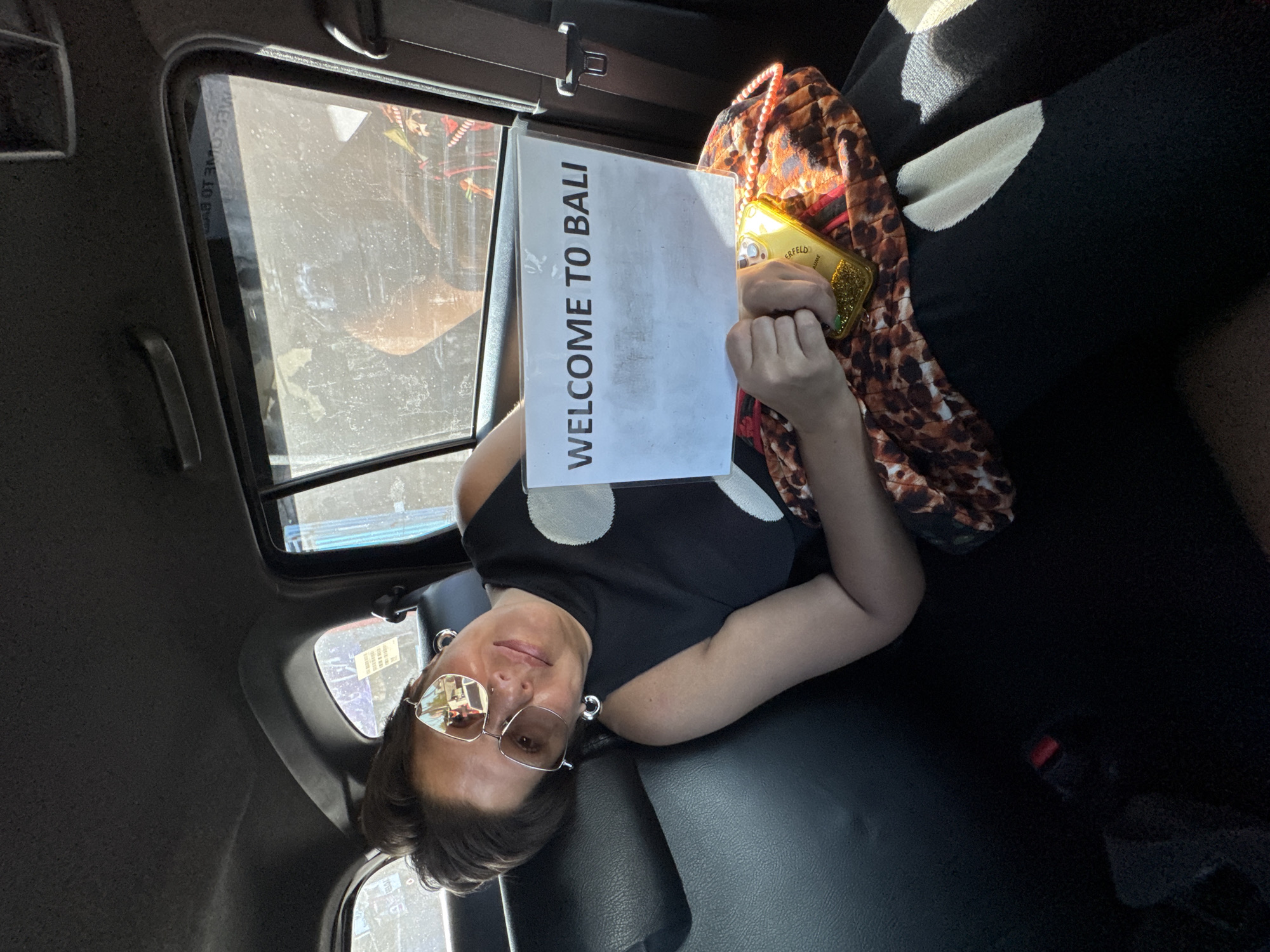 A person is seated in the back of a vehicle, holding a sign that reads "WELCOME TO BALI." The person is wearing glasses and earrings and is partially obscured by the sign. They are dressed in a black top with white polka dots and have a leopard-print bag beside them, along with a gold-colored phone case. The view through the car window shows a street scene, potentially reflecting urban scenery. The interior of the vehicle appears dark, with a black leather seat visible. It is possible that the location is in the city of Kuta Selatan, Indonesia, but this cannot be definitively confirmed without further information about the surrounding environment seen through the window.