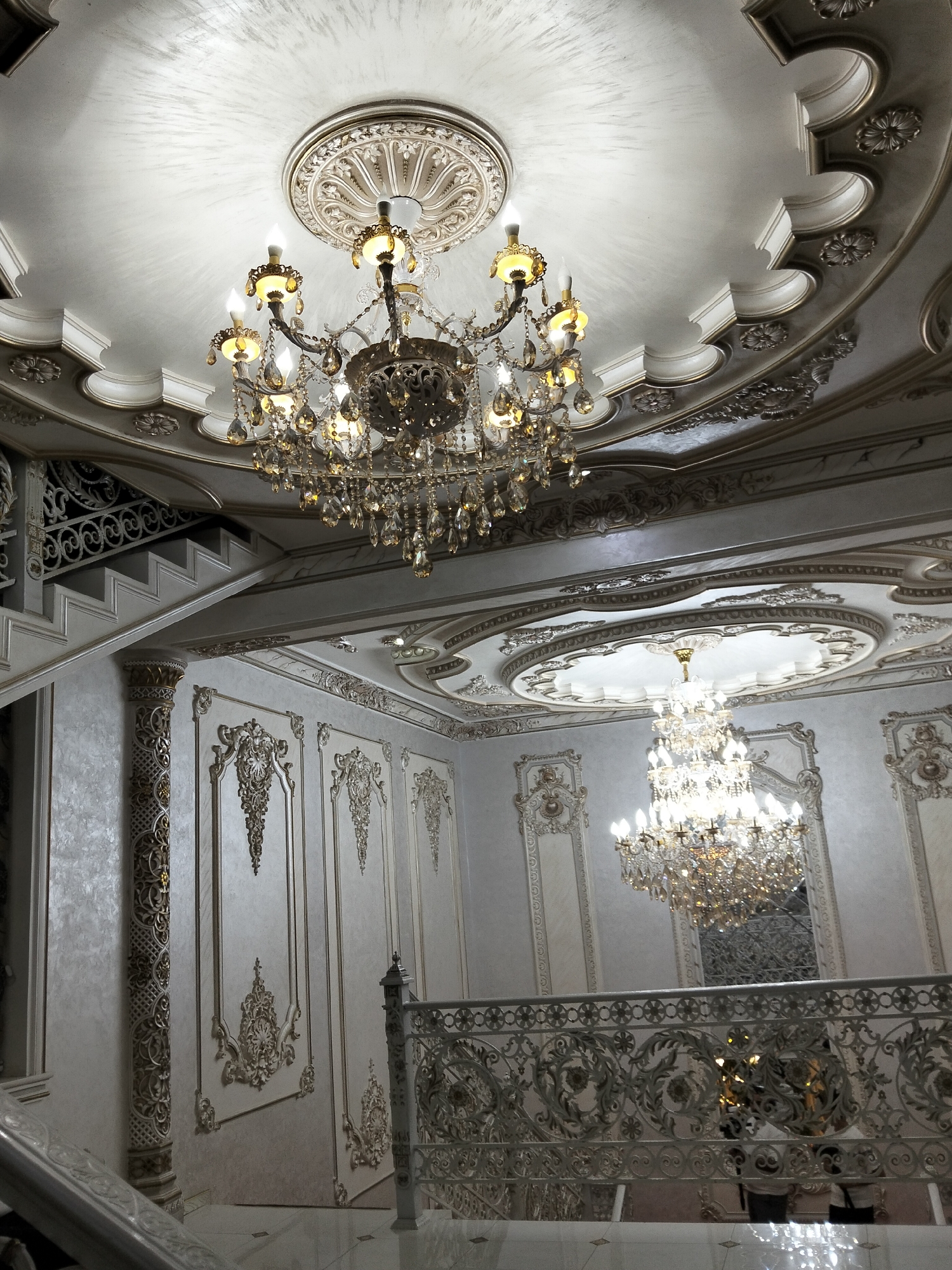 The image depicts an opulent interior space, likely a grand hall or a luxurious residence, with ornate architecture and elaborate lighting. Two large crystal chandeliers are prominently featured, one hanging from a decorated ceiling medallion and the other suspended from a more complex, multi-tiered ceiling design. The walls are adorned with intricate relief moldings and decorative paneling in shades of white and gold. A grand staircase with decorative metal railings is visible, ascending to an upper level. The flooring appears to be polished marble or tile. The overall ambiance is one of extreme wealth and detailed craftsmanship. The location in Kagan, Uzbekistan, suggests a context within a historically significant or culturally rich region, potentially a palace, a museum, or a high-end hotel. The scene is static and shows no people or overt activity.