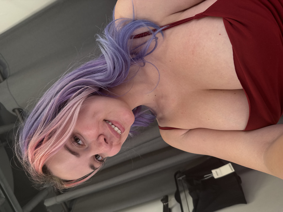 A young adult female is depicted in a close-up, chest-up shot within an indoor setting, identified as the City of Zagreb, Croatia. She has long, wavy hair, prominently dyed with pink/light reddish streaks at the front and purple/lavender for the majority. Her complexion is fair, and she is smiling directly at the camera, revealing teeth. She is wearing a red spaghetti-strap top or dress with a low neckline, exposing her upper chest and cleavage. The background features dark grey fabric, consistent with curtains or drapes, suggesting a fitting room environment. To the left, a dark garment with a white tag is partially visible, hanging on a rack or hanger.