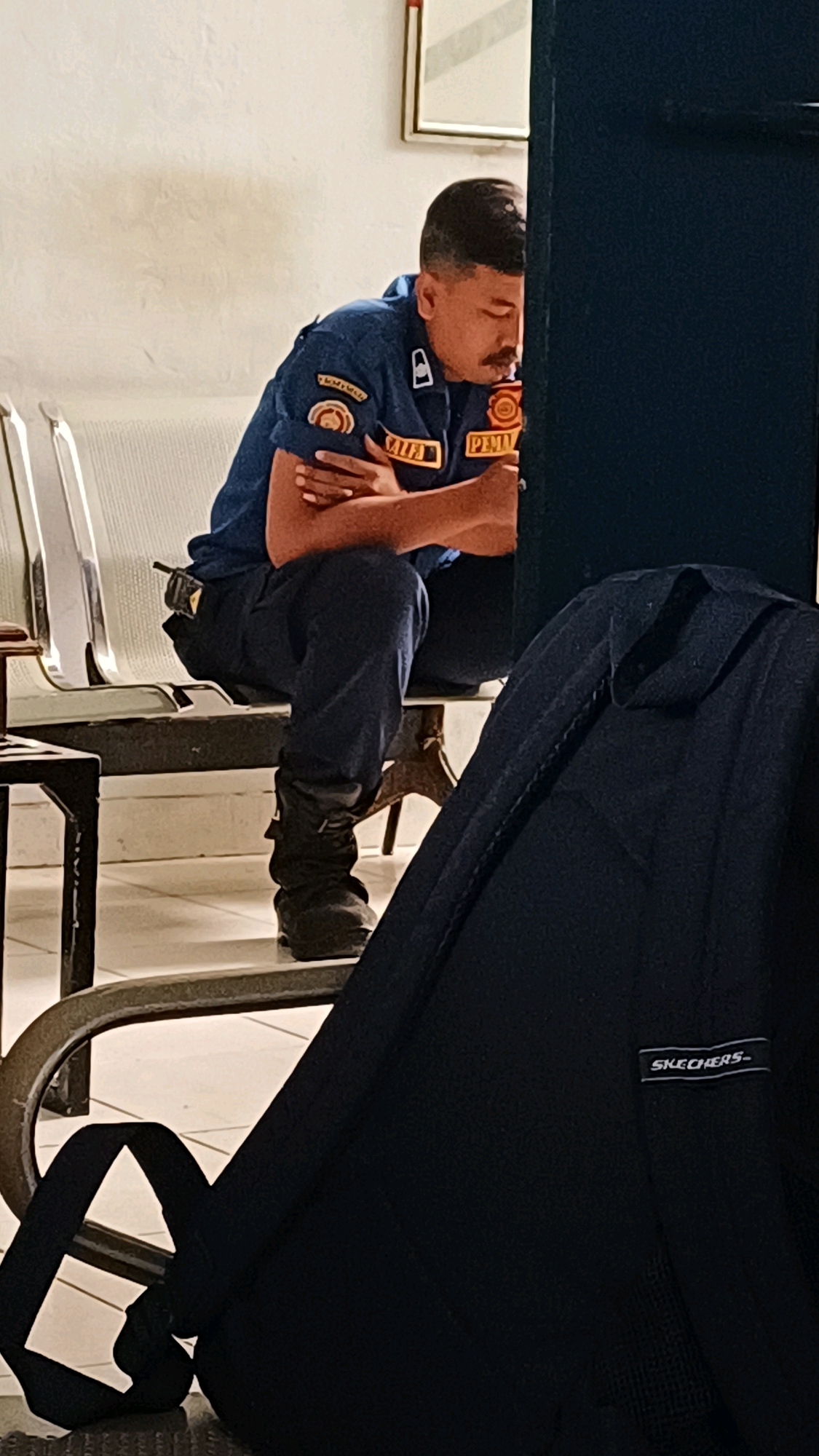 A man in a blue uniform sits on a metal bench with his arms crossed. He has a mustache and his eyes are closed. The uniform has patches on the sleeves and a name tag that reads "PEMA". He is wearing dark pants and black boots. A black backpack is placed on the floor in front of him. The background appears to be an indoor waiting area with a white wall and a framed picture. The image is likely taken in South Jakarta, Indonesia, given the context provided.