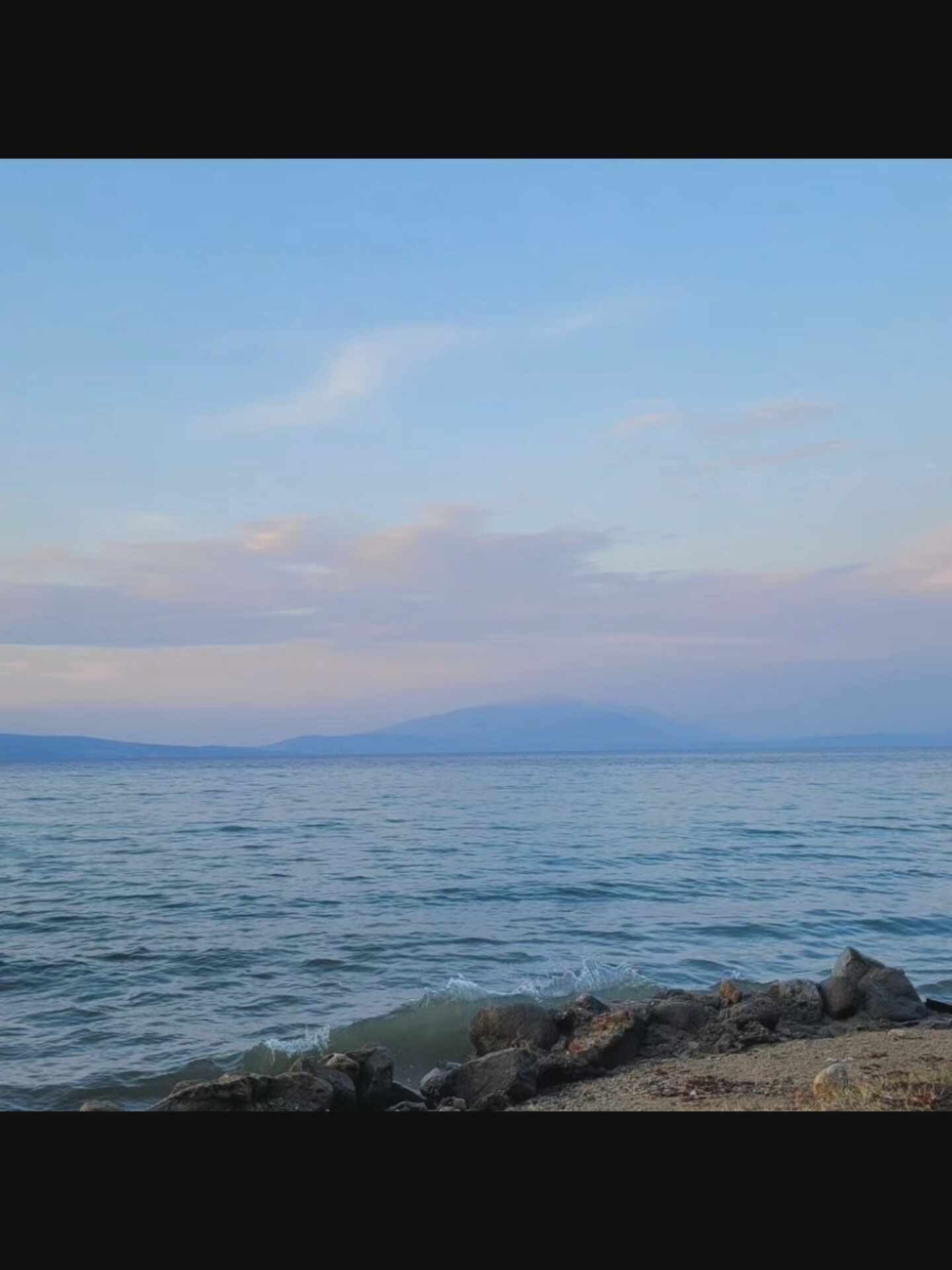 The image depicts a serene coastal landscape under a soft, pastel sky. The foreground features a rocky shoreline with small waves gently lapping at the stones. Beyond the immediate shore, a vast expanse of calm water stretches towards a distant, hazy mountain range. The sky is a gradient of pale blue and soft pinks and purples, suggesting either dawn or dusk. There are no people or man-made structures visible in the scene. The overall impression is one of natural tranquility. The location is identified as Bielawska Wieś, Poland, a detail that does not provide specific visual cues within the image itself.