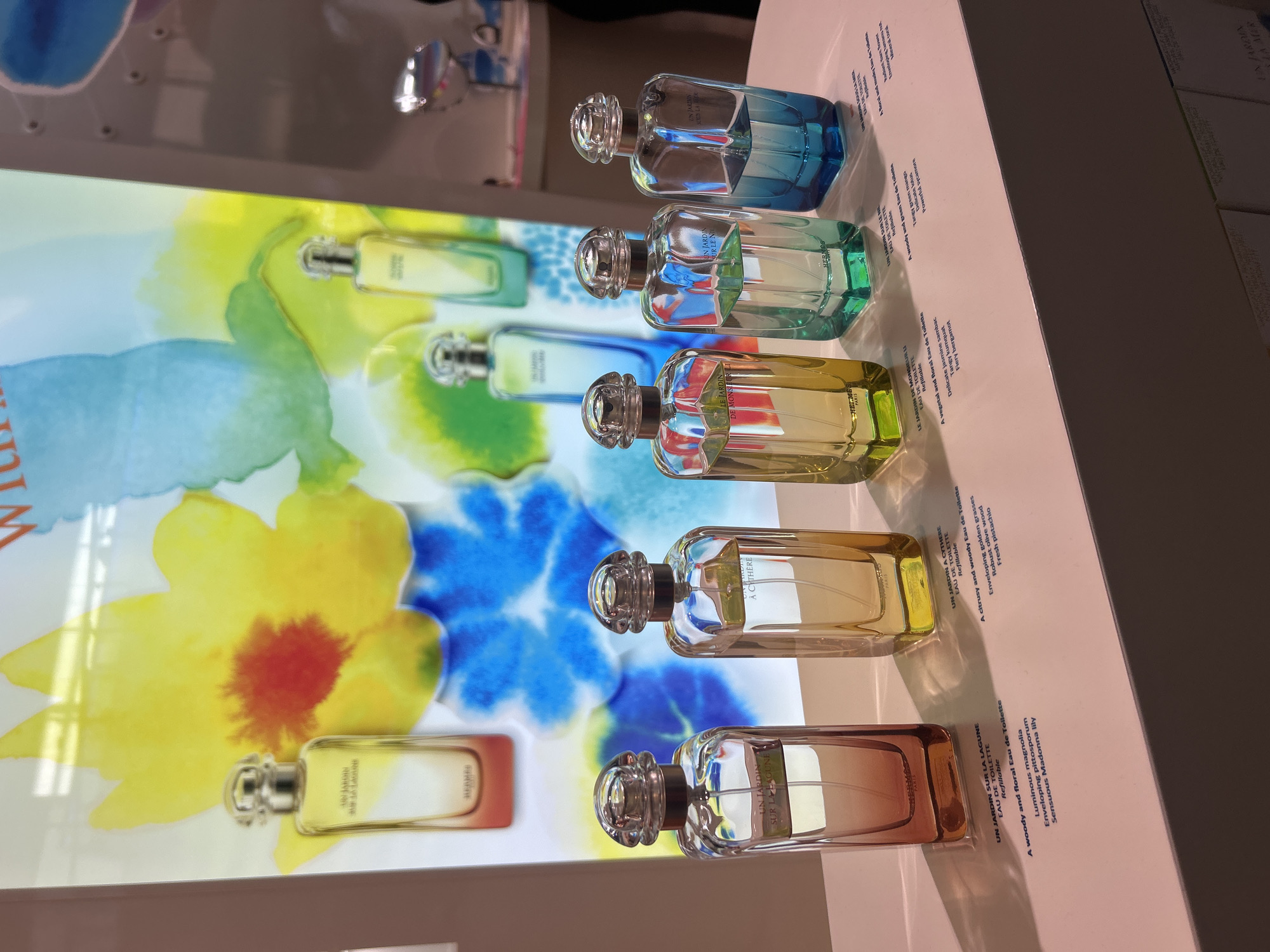The image displays a retail display of Hermes perfumes in Tambon Mai Khao, Thailand. Five perfume bottles are arranged in a row on a pink surface. The bottles are clear glass with silver caps, and the liquid inside varies in color from orange-red to blue. The background features a large, abstract floral watercolor painting with yellow, red, blue, and green tones. Text descriptions of the perfumes are visible on the pink surface below the bottles. There are no visible people in the image. The lighting is bright, suggesting an indoor retail environment.
