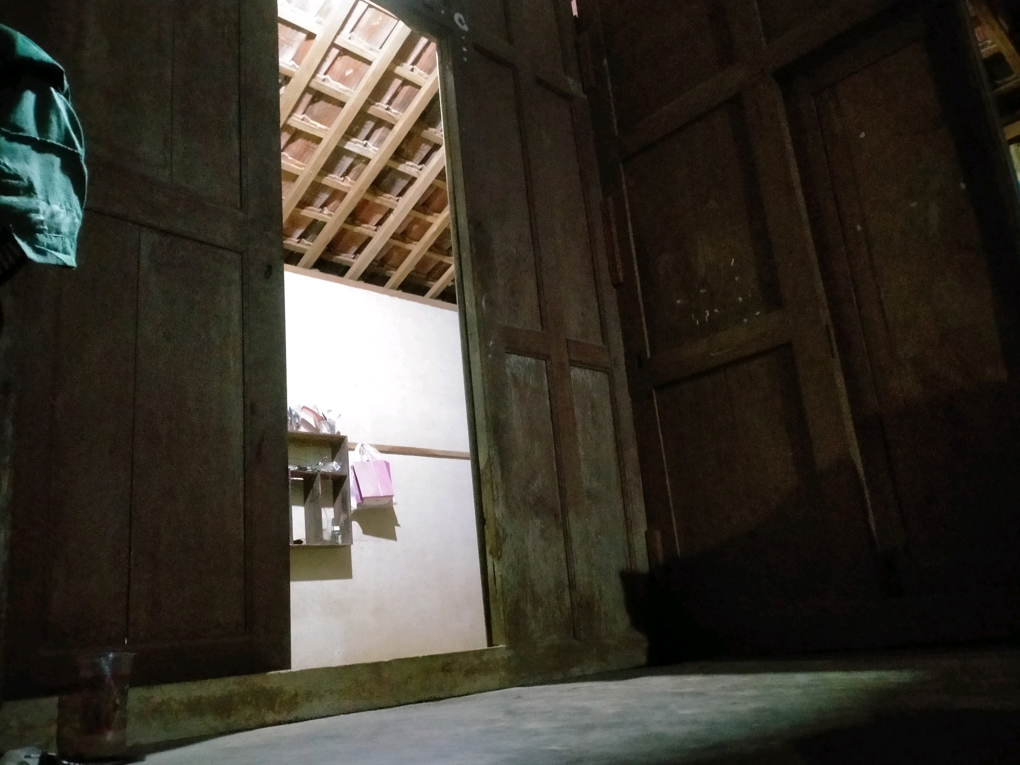 The image depicts an interior view of a rustic dwelling, likely a house, located in Playen, Indonesia. The foreground is dominated by dark, weathered wooden doors, with one door ajar, revealing a glimpse into another room. The lighting is dim, with a yellowish hue emanating from an unseen source.
The opened door leads to a space with a light-colored wall. On this wall, a simple wooden shelf unit is mounted, holding a few items. Two pink shopping bags hang from the shelf. Above this area, the exposed wooden rafters of the ceiling are visible, covered with what appear to be clay roof tiles, characteristic of traditional Indonesian construction.
To the left of the main doorway, a green garment, possibly a jacket, is hanging on the edge of the door. In the lower left corner, a partially visible glass contains a dark liquid. The floor in the foreground is concrete, appearing somewhat grimy. The right side of the image is largely obscured by shadow, suggesting another room or alcove, with the dark silhouette of what could be a doorway or furniture.
The overall impression is of a simple, possibly older, residential structure, with visible elements of traditional building materials and a somewhat cluttered, lived-in atmosphere. The location in Playen, Indonesia, implies a rural or semi-rural setting.