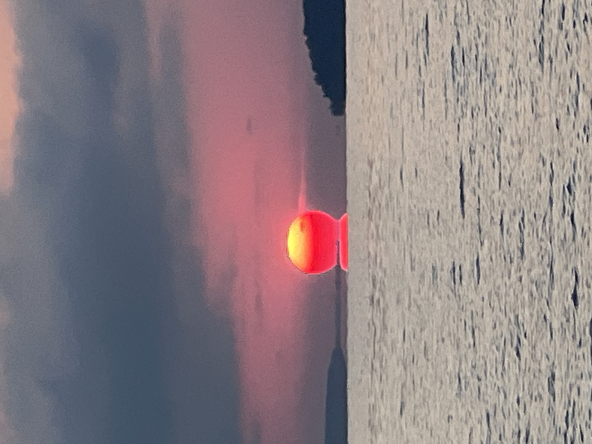 The image depicts a sunset over a body of water, likely a lake or sea, with a prominent sun in a bright orange and pink hue. The sun appears almost perfectly spherical and is low on the horizon, creating a reflection on the water. Dark clouds dominate the upper portion of the sky, contrasting with the vibrant colors of the sunset. The water is calm with gentle ripples. On the left and right sides of the frame, distant landmasses are visible as dark silhouettes against the lighter sky. The scene is devoid of any human presence or man-made structures, suggesting a natural, undisturbed landscape. The image is a photograph, possibly taken during dusk. The location is stated to be Novopichugovo, Russia.