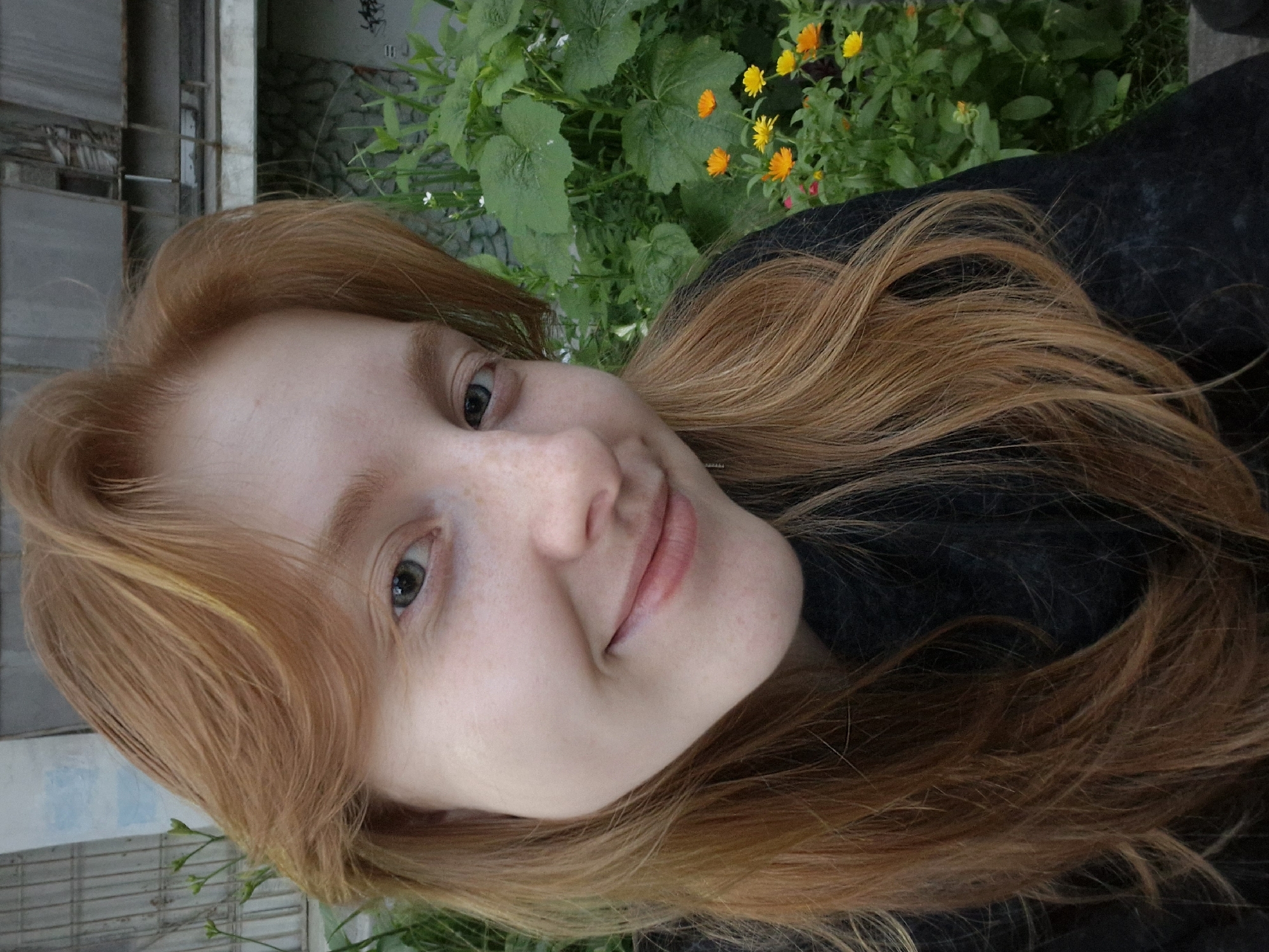 This is a vertical, close-up photograph of a young woman with red hair. She is outdoors, with green foliage and small yellow and orange flowers visible in the background, suggesting a garden or natural setting. The woman's head is tilted, and she is looking towards the camera with a slight smile. Her skin has a fair complexion with freckles. Her hair is long and wavy, falling around her face and shoulders. She is wearing a dark, possibly black, garment. The lighting suggests it might be daytime, possibly overcast or late afternoon given the soft light. There is no visible text in the image. The context places this scene in Yekaterinburg, Russia.