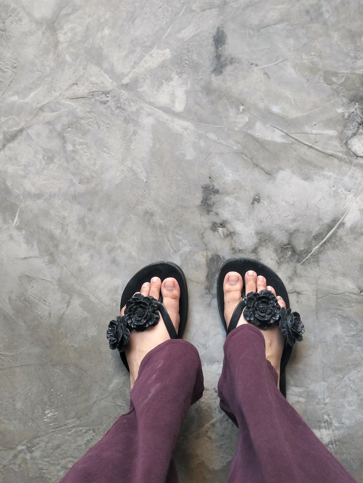 A pair of feet wearing black flip-flops with floral embellishments and dark maroon pants stand on a textured concrete floor. The image appears to be taken from a high angle, looking down at the feet and the floor. The concrete floor exhibits variations in color and texture, with visible cracks and subtle shading. The lighting suggests an indoor setting, possibly a studio or a room with overcast natural light. The location is specified as Caoayan, Philippines.