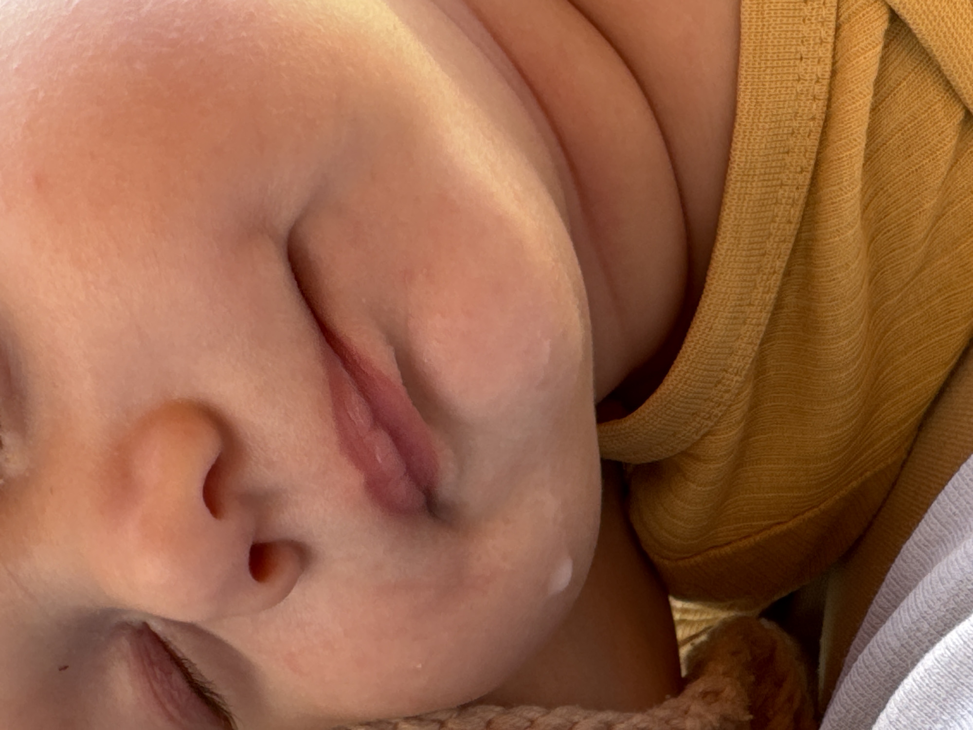 The image is a close-up, overhead shot of a sleeping baby's face. The baby's eyes are closed, and their mouth is slightly open, with the lips appearing soft and pink. The baby's skin is smooth and fair. The baby is wearing a mustard-yellow, ribbed garment, possibly a onesie or shirt, which is visible on the right side of the frame. The garment has a textured weave. A light-colored, woven fabric, perhaps a blanket or crib lining, is visible at the bottom of the image. The lighting suggests it could be daytime, with a soft, natural light illuminating the baby's face. The overall mood of the image is peaceful and intimate, capturing a moment of restful sleep. No text is visible in the image. The location context of Патрушева, Russia, does not provide specific visual cues within this close-up image.