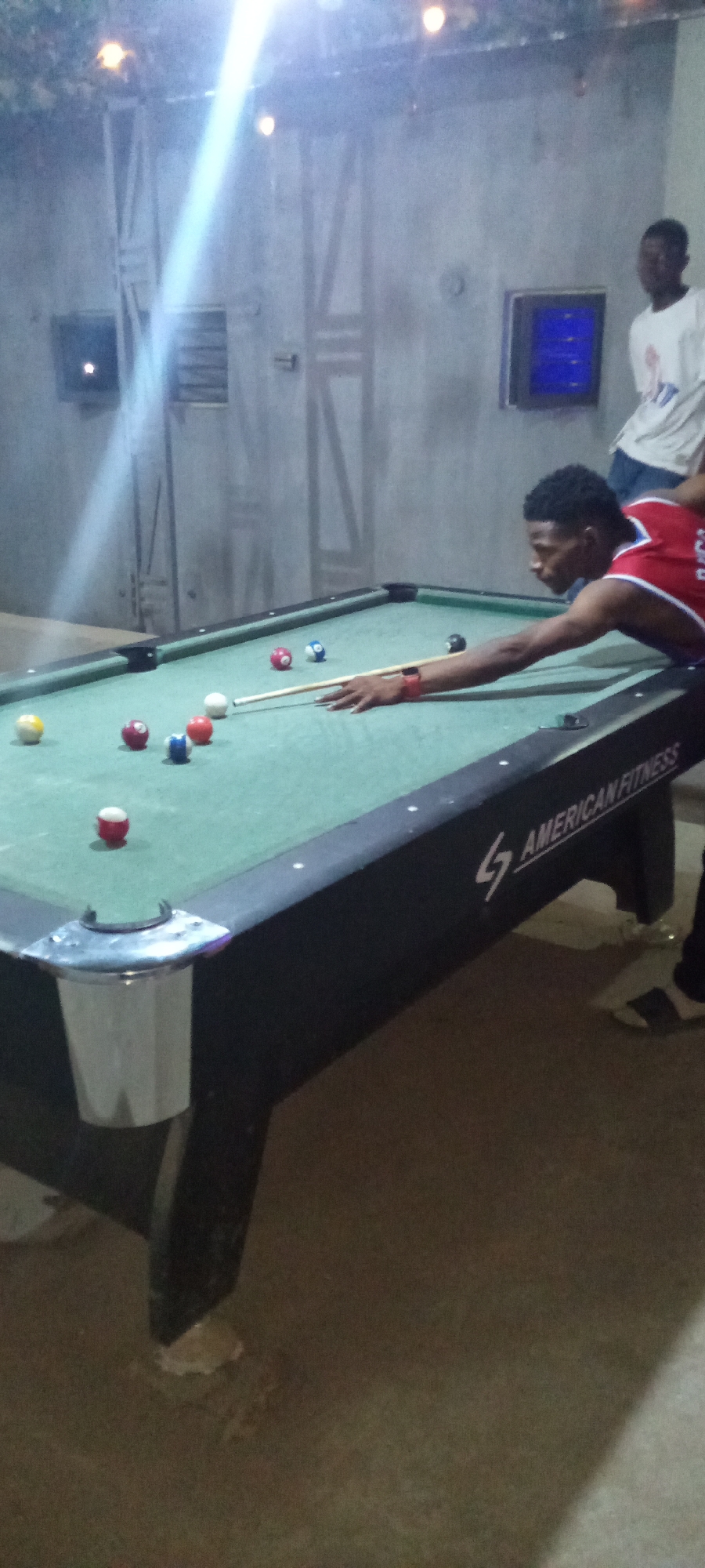 The image depicts an indoor scene in Akwanga, Nigeria, where two young men are playing or observing a game of pool, likely in the evening or at night.
The main subject is a young man, possibly in his late teens or early twenties, positioned centrally and leaning over a green-felted pool table. He is actively aiming a cue stick at the white cue ball, preparing for a shot. He wears a red and blue sleeveless top, resembling a sports jersey, and has a red wristband on his left arm. Various colored pool balls, including solids and stripes, are scattered across the table's surface.
In the background, another young man, partially visible and somewhat blurred, stands near a light-colored wall, wearing a white t-shirt. The room's walls appear to be concrete or painted a light gray, featuring industrial-style panels or structures, some with embedded blue light. Overhead, several small, warm-toned lights are visible, along with a prominent bright light source from the upper left casting a strong beam across the ceiling and wall.
A notable detail is the text "AMERICAN FITNESS" displayed in white on the dark side panel of the pool table, suggesting the brand or a venue name. The overall ambiance is dim, indicating an indoor recreational setting. The player's posture conveys focus and concentration on the game.