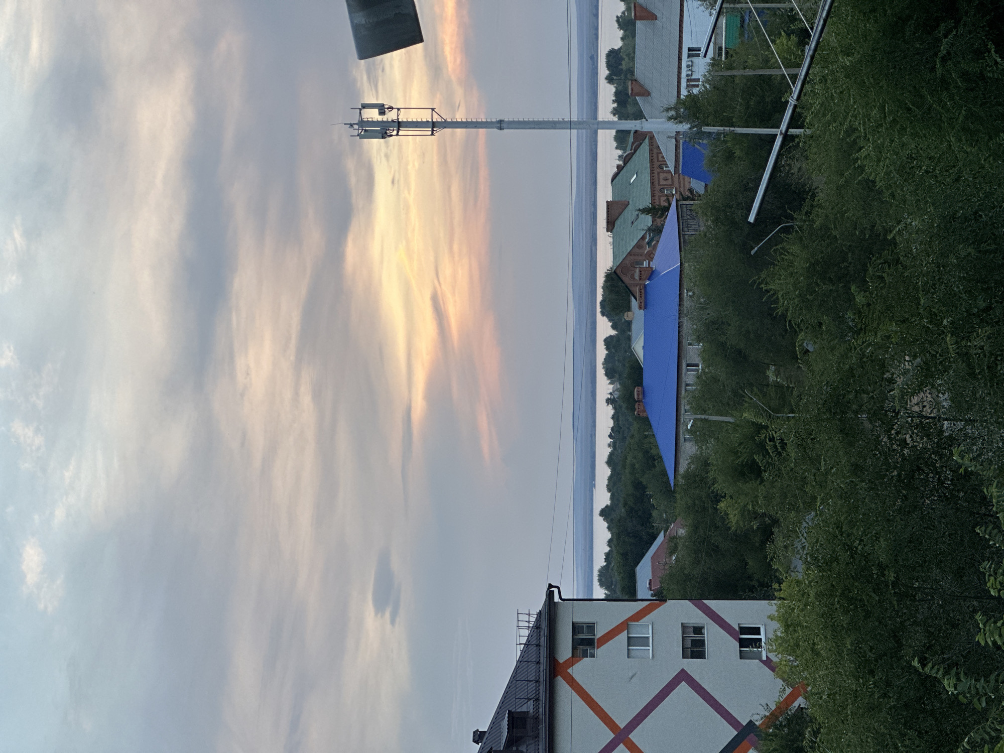 The image depicts a view of the city of Energetik, Russia, from an elevated position. In the foreground, dense green foliage obscures much of the lower part of the scene. A multi-story apartment building with a dark roof and a white facade decorated with colorful geometric lines is visible on the left. Several houses with various roof colors, including blue and green, are scattered throughout the mid-ground, surrounded by trees. A tall cell tower stands prominently in the mid-right of the frame. Beyond the city, a wide body of water, likely a lake or river, stretches across the horizon, bordered by a line of trees. The sky above is filled with soft, wispy clouds, illuminated by the warm hues of either a sunrise or sunset, suggesting a twilight setting. There are no people visible in the image.