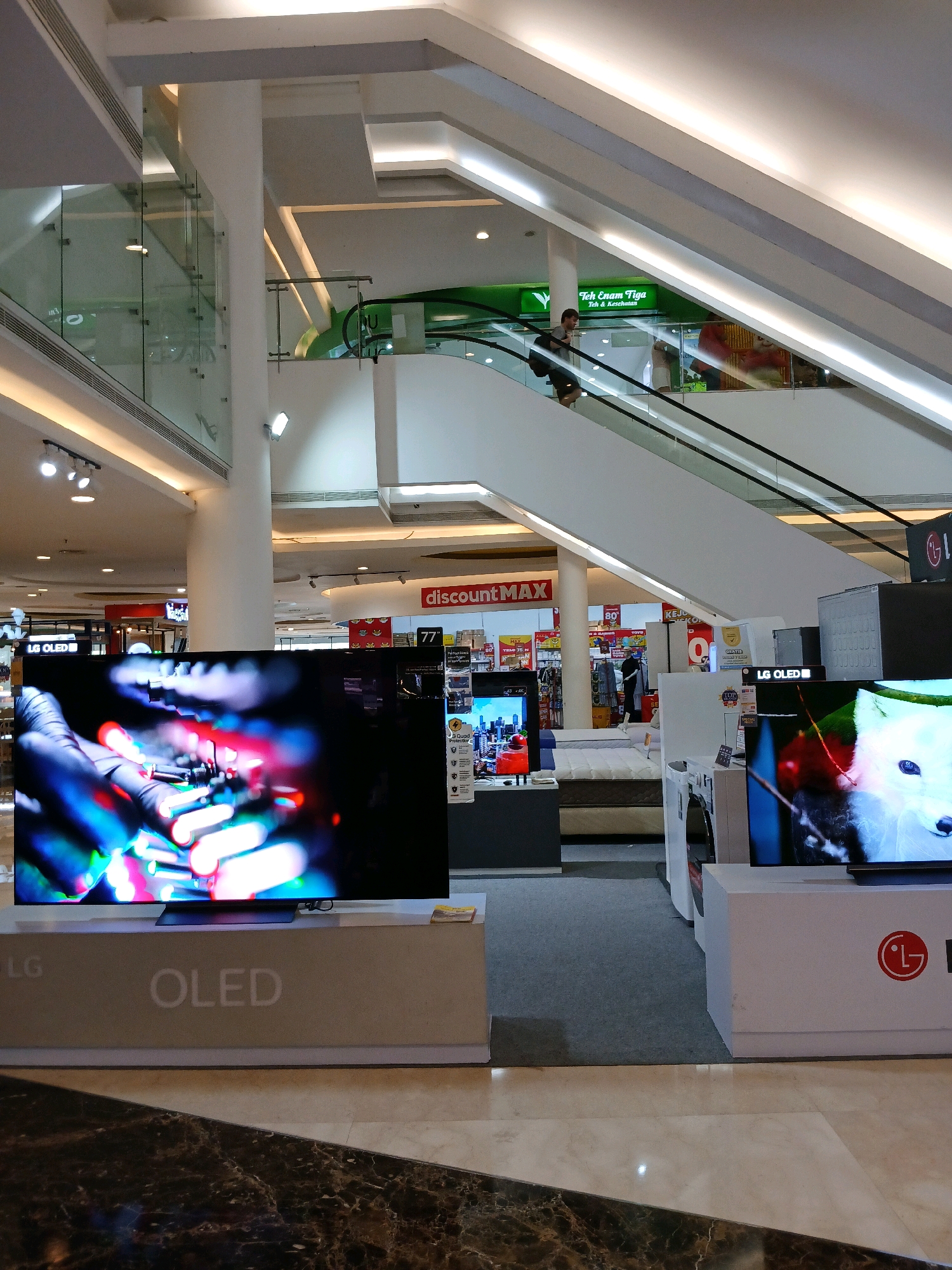 An interior shot of a large electronics store in the city of North Jakarta, Indonesia, focuses on several large LG OLED televisions displayed on white stands. One television in the foreground displays a close-up image of colorful lights. The background shows an escalator with a few shoppers, an advertisement for "Teh Enam Tiga" (Six Three Tea), and various other electronic appliances and displays. The store appears to be multi-leveled, with a modern architectural design featuring white pillars, glass railings, and bright overhead lighting.