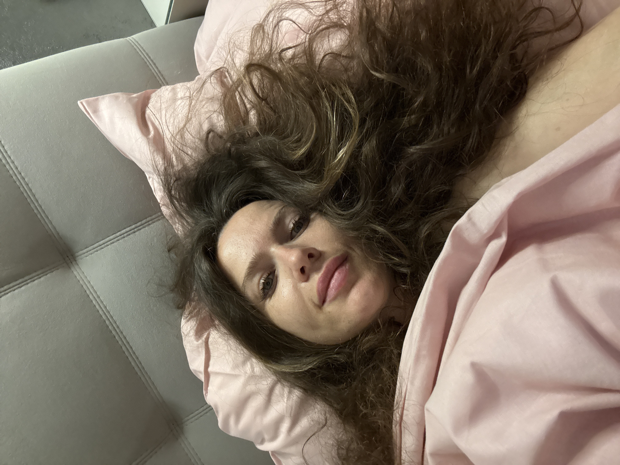 A medium close-up shot captures a woman lying on a bed, looking directly at the camera. She appears to be in her 20s or 30s, with long, wavy, dark brown hair spread out around her head on a light pink pillow. Her face is in the center of the frame, showing her open eyes, a slightly parted mouth with full lips, and a neutral, contemplative expression.
She is covered partly by a light pink blanket or duvet, matching the pillowcase. To her left, a grey, padded headboard or bed frame with horizontal stitching lines is visible. The background beyond the headboard is indistinct. The lighting is soft and even, suggesting an indoor setting. The image portrays a moment of repose or casual interaction, likely in a bedroom. The location context provided is Moscow, Russia. There is no visible text in the image.