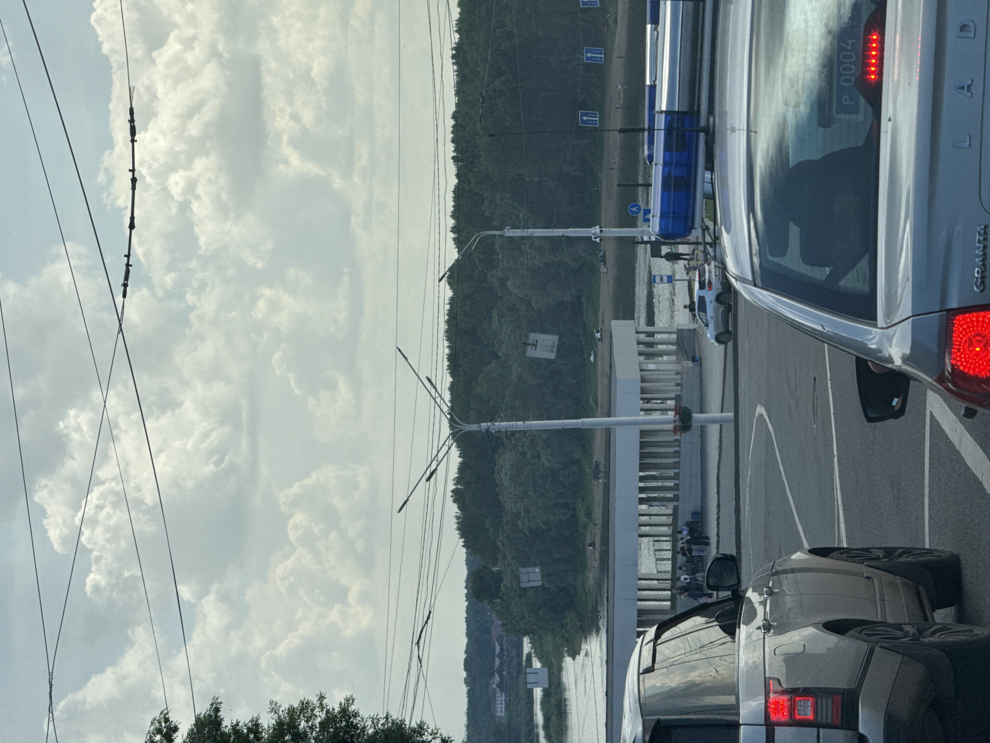 A high-angle medium shot captures a traffic scene with vehicles and a waterfront environment. In the foreground, the rear of a silver SUV is visible, showing its taillights and a license plate that reads "P 0004". To the right, another car's taillight is also lit. Beyond the vehicles, a wide river or body of water separates the foreground from a densely wooded shoreline. Several people are gathered on the bank near the water, and a few cars are parked or driving along the shoreline road. A modern building with a colonnade runs along the waterfront in the middle ground. Overhead, multiple power lines crisscross the cloudy sky. The scene appears to be set in a city, likely during daylight hours, with a blend of urban infrastructure and natural scenery.