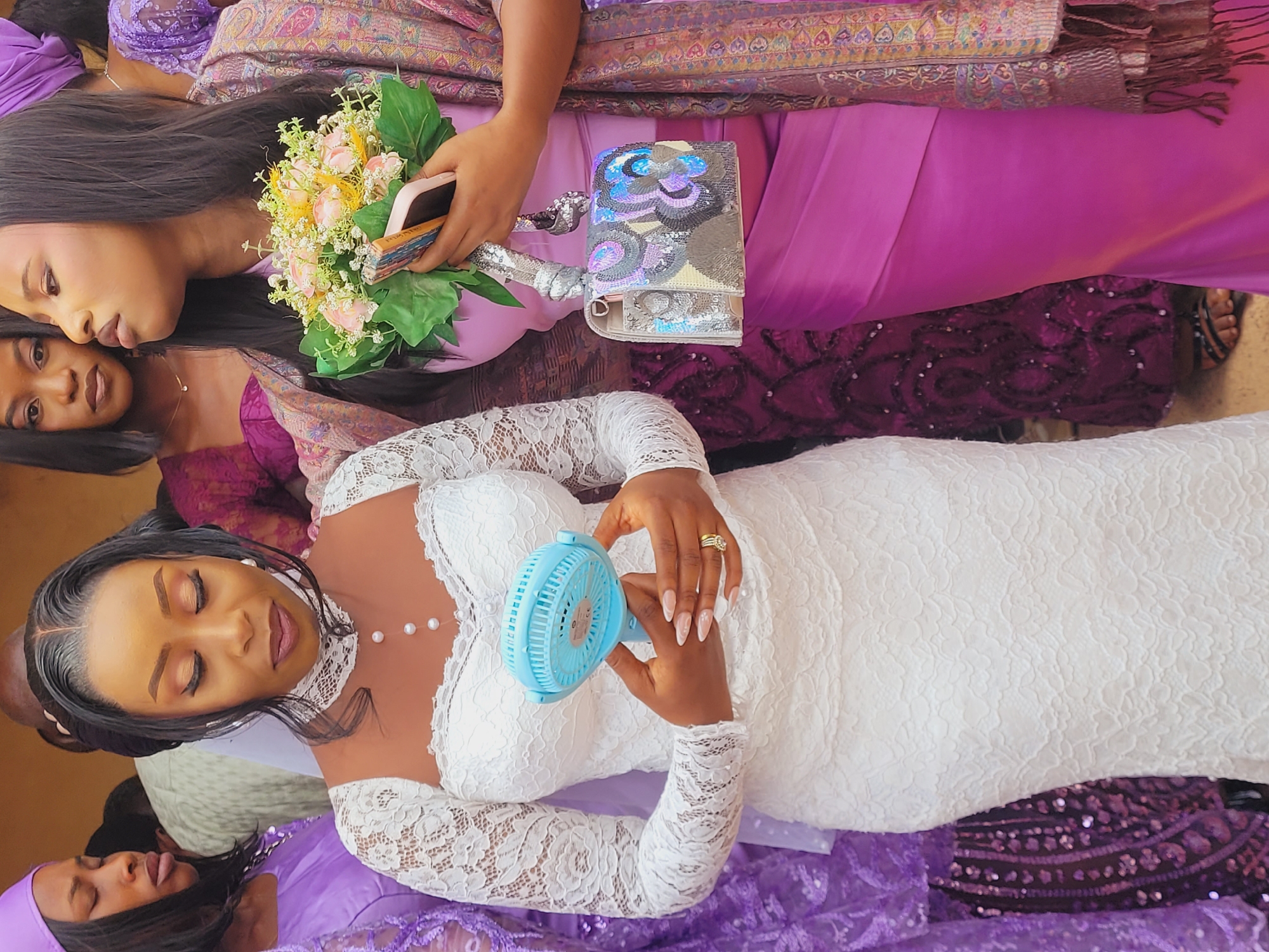 The scene is located in Bukuru, Nigeria. A dark-skinned woman with dark hair, styled with a center part and framing strands, is centrally positioned, her eyes downcast. She wears a white lace long-sleeved dress featuring a sweetheart neckline and a sheer high collar adorned with small pearl buttons. A multi-band gold ring is on her left ring finger, and her nails are long, pointed, and manicured with a light polish. She holds a light blue portable mini-fan with both hands at her midsection.
To her immediate right, a dark-skinned woman with long, straight dark hair, partially obscured, wears a pink top and skirt ensemble with a multicolored patterned shawl (purple, pink, gold) draped over her shoulders. She holds a small bouquet of artificial light pink and yellow flowers along with a light-colored smartphone in her left hand. A silver-toned rectangular clutch purse with abstract sequin patterns in blue, purple, grey, and white hangs from her left hand by a sequined strap.
To the far left, a third dark-skinned woman in a purple headwrap and purple lace garment is partially visible, also with downcast eyes. Several other blurred figures, predominantly women in purple or maroon attire, are visible in the background, indicating a gathering. The setting appears to be an indoor or semi-enclosed area with warm-toned walls and bright, natural light.
