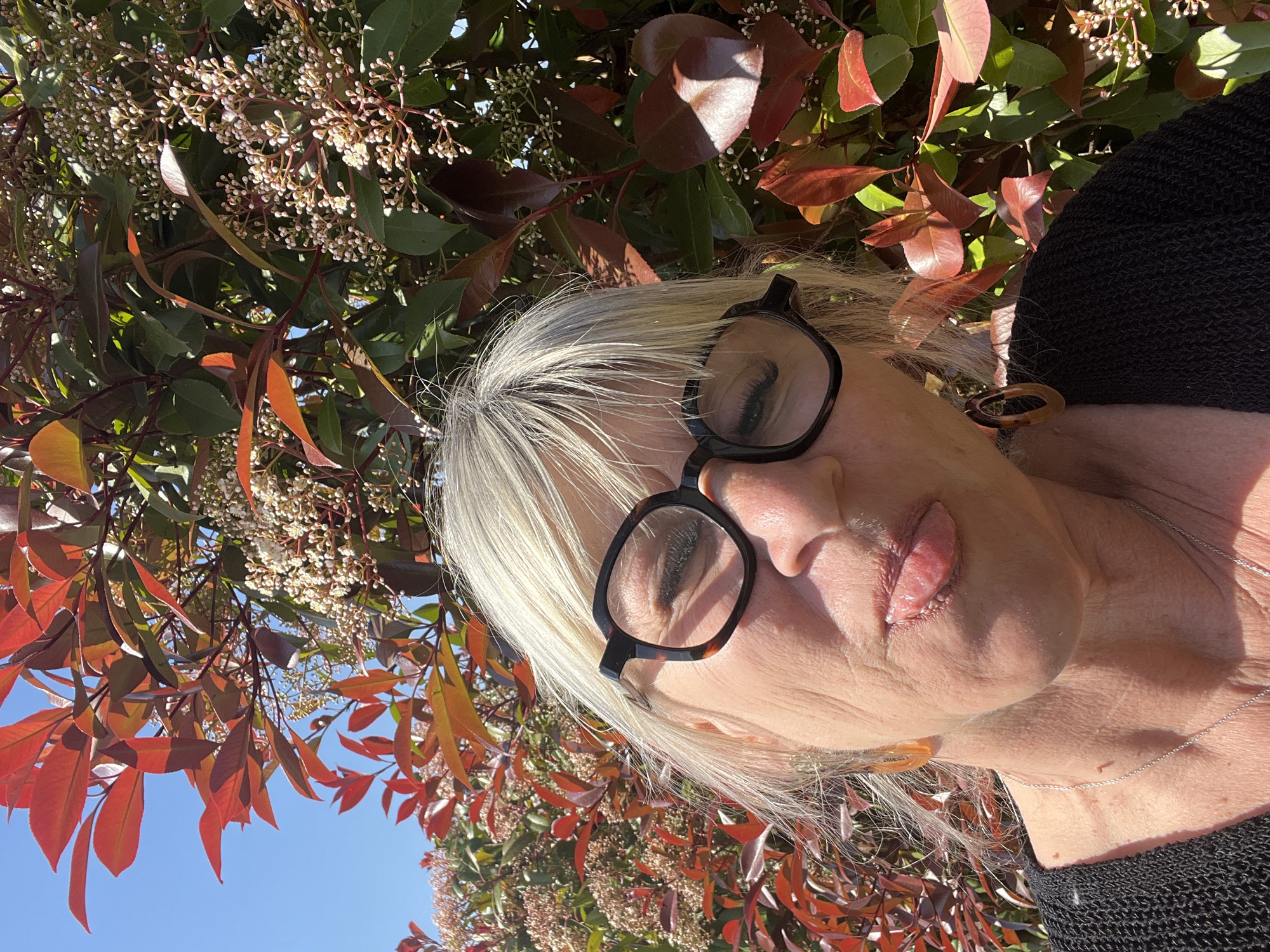 The image is a close-up, tilted shot of an older Caucasian woman with closed eyes, wearing black-rimmed glasses. She has silver hair and a black, knit top. A silver necklace is visible around her neck. The background consists of foliage, including leaves that are red and green, and clusters of small, white flowers. The scene is outside, as evidenced by the clear blue sky visible through the leaves. There is no information to suggest this image was taken in Chambœuf, France.