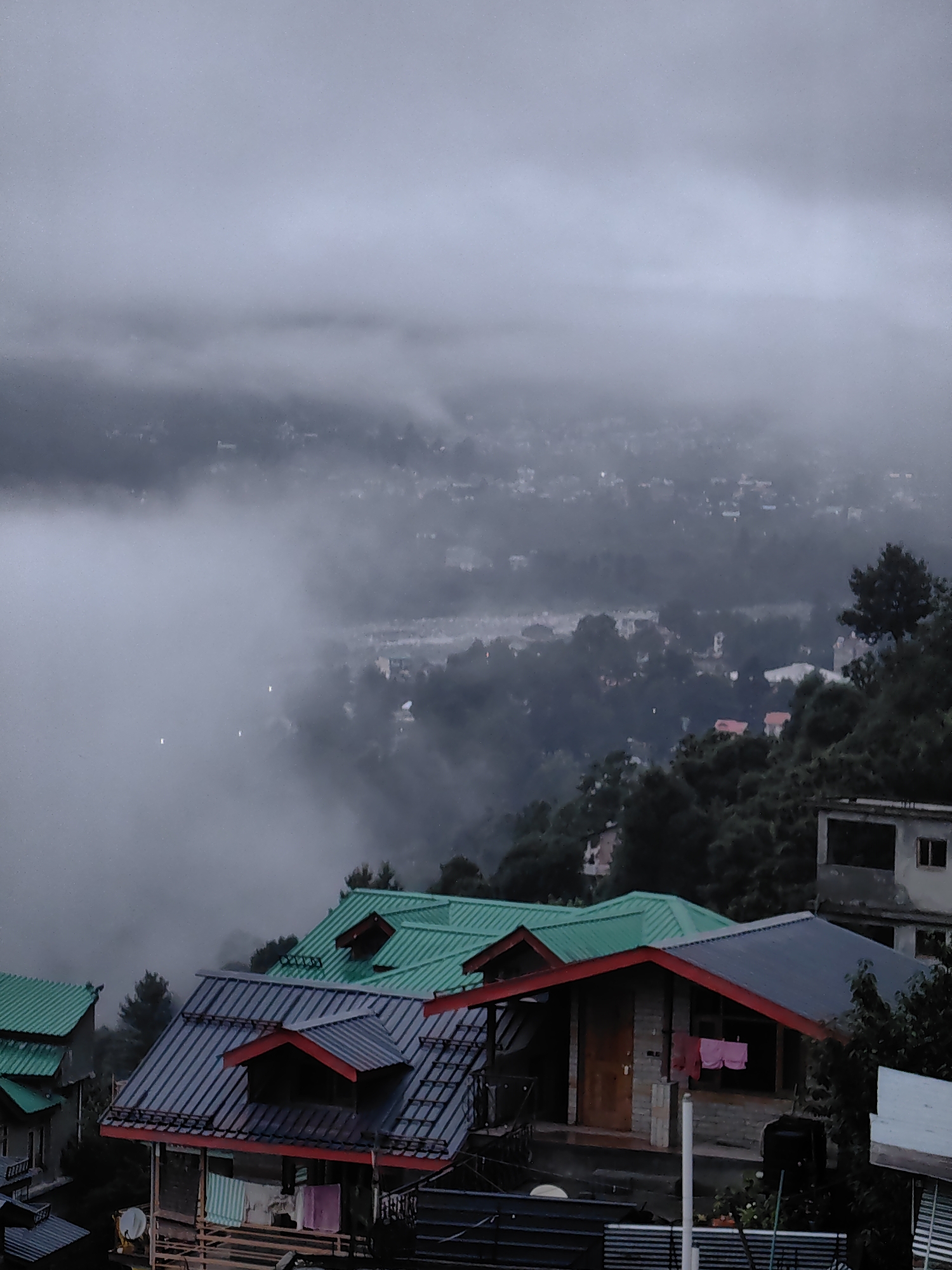 The image depicts a misty view of houses in the city of Vashisht, India, under a cloudy sky. In the foreground, several houses with sloped, multi-colored roofs are visible. One house features a dark gray roof with red trim and a wooden door, with pink shorts hanging on a clothesline outside. Another house has a bright green roof with two dormer windows. In the background, the city sprawls across a hillside, partially obscured by thick fog and mist, with more buildings and trees discernible through the haze. The overall scene conveys a cool, damp atmosphere, characteristic of a cloudy day in a mountainous region.
