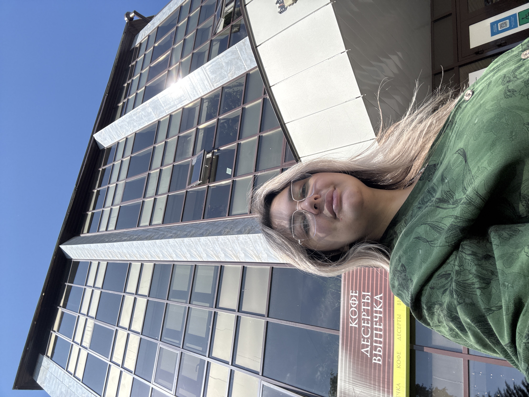 The image shows a young woman with blonde hair and glasses, wearing a green patterned jacket. She is standing in front of a modern building with large glass windows. The building has a sign that reads "КОФЕ ДЕСЕРТЫ ВЫПЕЧКА", which translates to "Coffee Desserts Baking". The sun is shining brightly, casting reflections on the windows. The image appears to be taken in Birobidzhan, Russia, given the Cyrillic text on the sign. The overall scene is bright and urban, with the focus on the woman and the building.