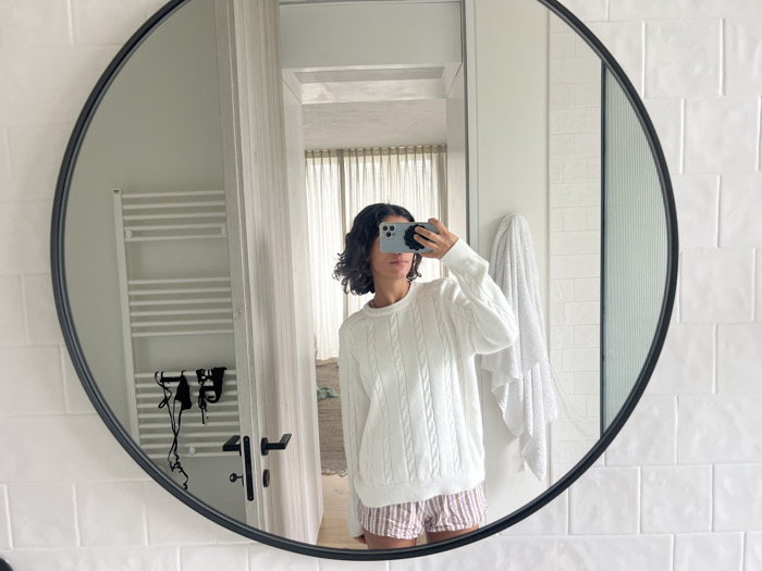 The image presents a mirror selfie taken in a bathroom located in Vila Nova de Cerveira, Portugal. A circular mirror with a black frame reflects a person, whose face is partially obscured by a light-colored smartphone held horizontally to take the photograph. The individual wears a white, cable-knit long-sleeved sweater and light-colored, striped shorts. Visible in the reflection behind the subject is a white tiled wall. To the left, a white, horizontally-barred towel radiator is mounted, with a black hairdryer and its cord hanging below it. To the right, a white towel is draped over a fixture. An open doorway in the background reveals an adjacent room with light-colored flooring and a window featuring sheer curtains.