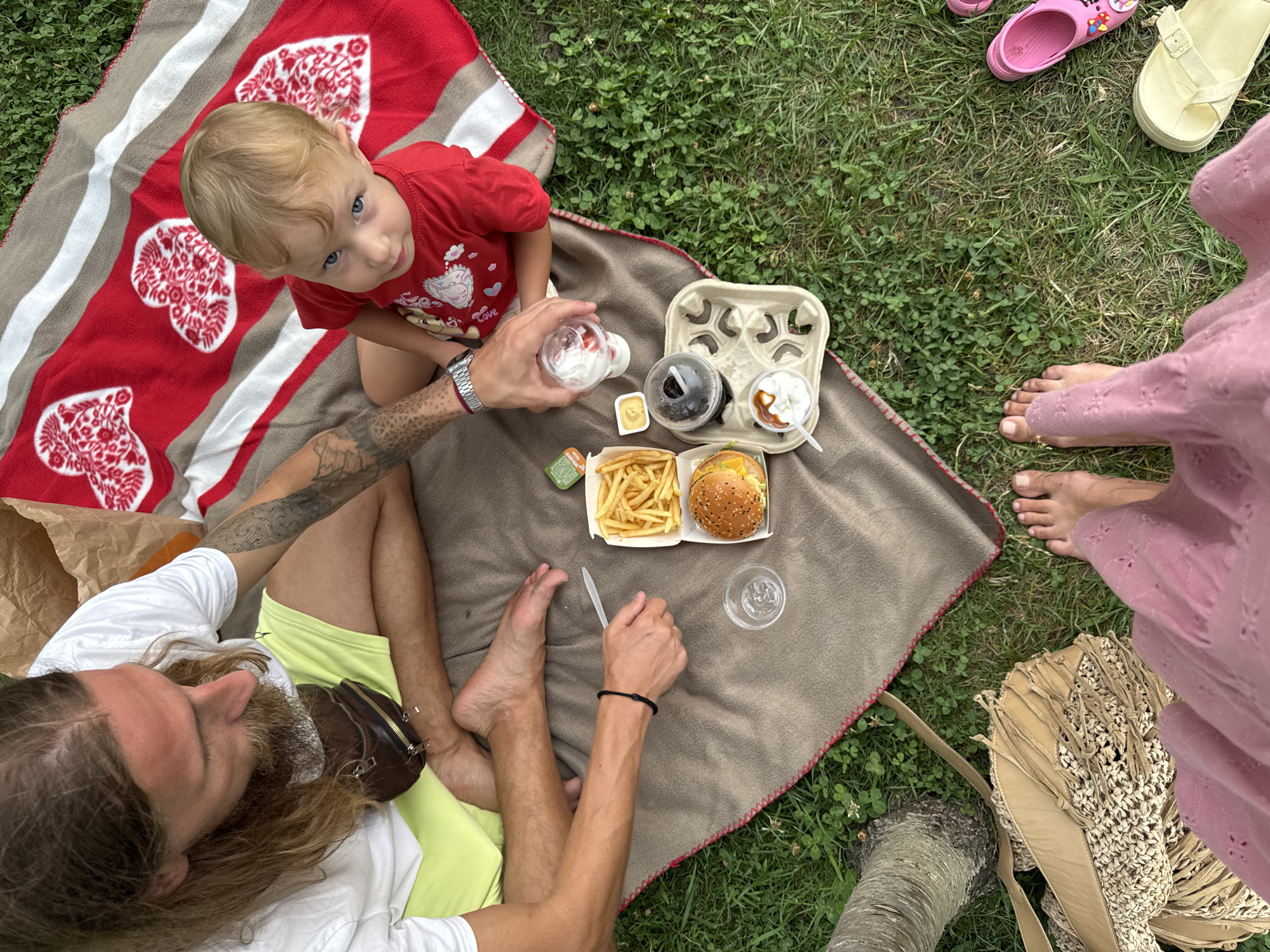 The image depicts a family picnic on a grassy area, likely in Sochi, Russia, given the provided context. A man with long hair and a beard, wearing a white t-shirt and yellow shorts, sits on a brown blanket, facing away from the camera. He has tattoos on his arm and is holding a white plastic utensil. Next to him, a young child with blue eyes, wearing a red t-shirt, sits on a red and white striped blanket adorned with heart patterns. The child is looking towards the camera. On the blanket, there is a picnic spread including a burger, french fries, a drink in a clear cup, and two desserts in cups. A brown paper bag is visible in the background. In the foreground, two pairs of bare feet are visible, one belonging to the man and another, presumably belonging to the viewer. A straw woven bag with fringes and a pink dress are also visible in the foreground. To the right, a pair of pink Crocs and yellow sandals are placed on the grass. The environment is grassy with scattered clover plants, suggesting an outdoor setting in a park or a similar recreational area.