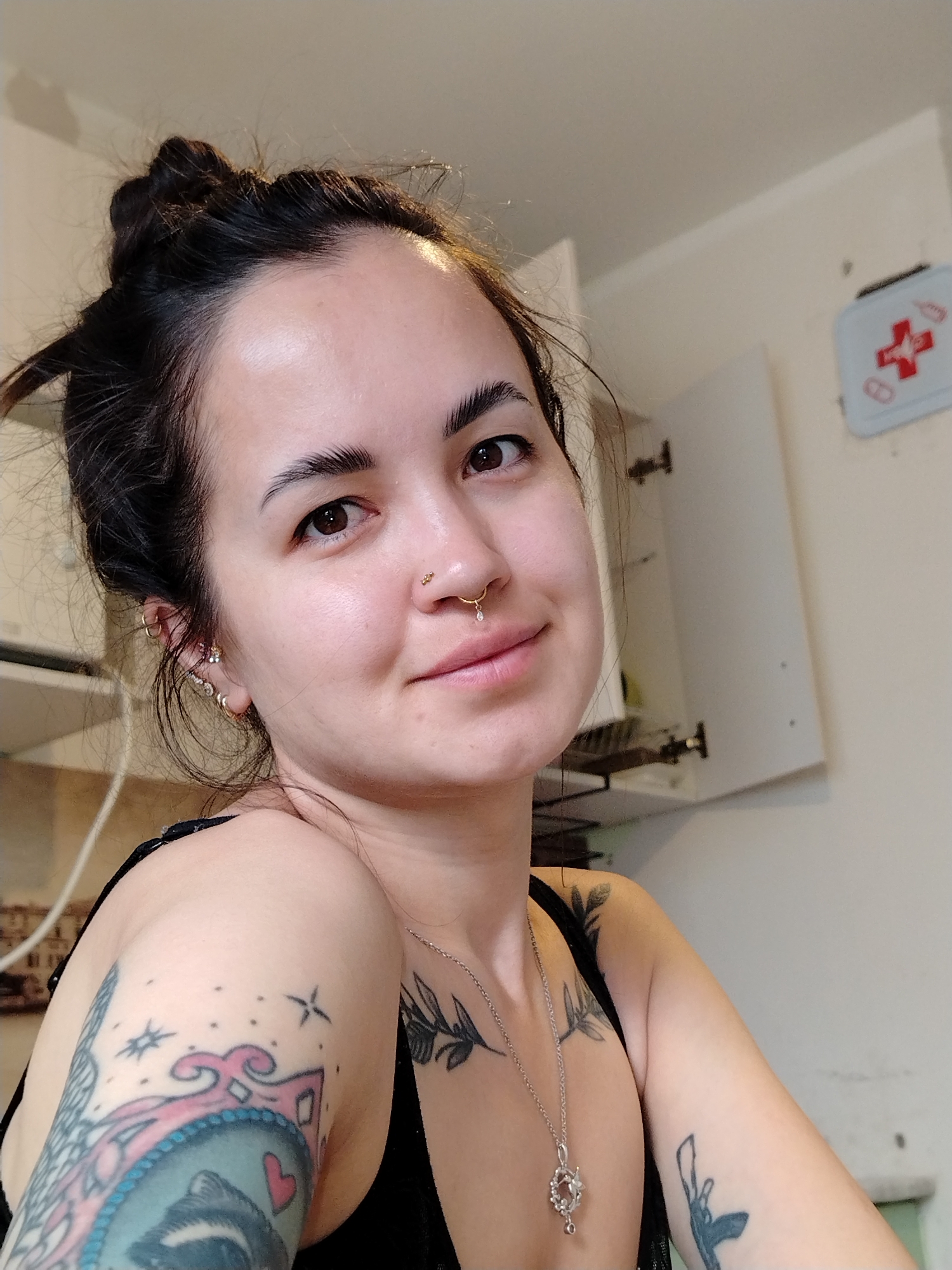 A young woman is in a kitchen. She has dark hair pulled up in a bun. She has multiple piercings in her ear and nose. She wears a black top and a silver necklace. She has several tattoos on her arms and chest. An open cabinet and a first aid kit are on the wall behind her. There is no indication of this being Noviy, Russia.