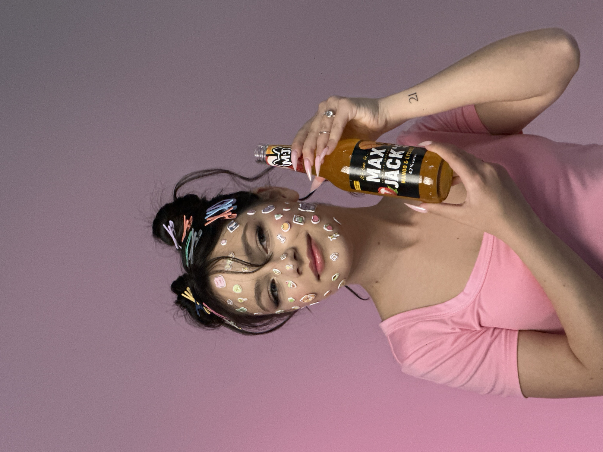 A woman is featured in the image, lying down with her face adorned with stickers and her hair held up with colorful clips. She's wearing a pink t-shirt and holding a bottle of "Max Jack Mango & Lychee" beverage. The backdrop is a gradient of pink hues. The woman has a tattoo on her arm displaying the number "21." The scene is not explicitly set in Yerevan, Armenia, nor are there any visible landmarks to ascertain the location.