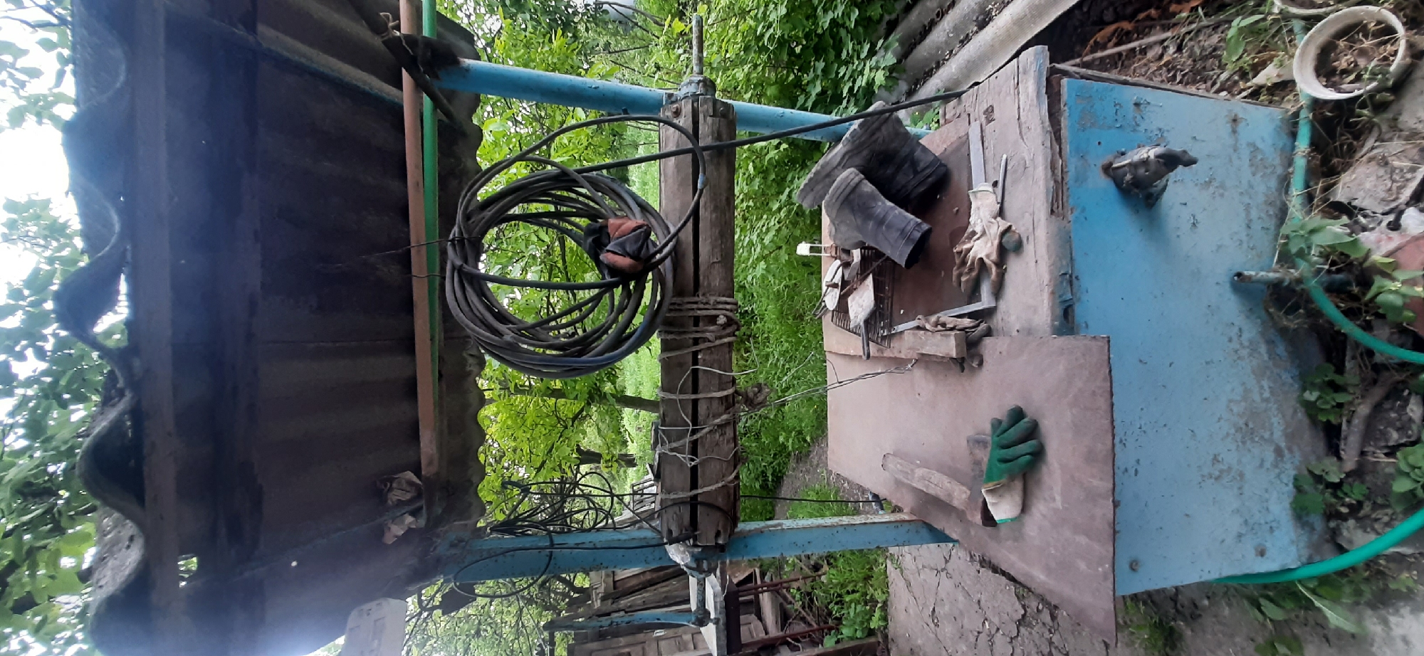 The image depicts an outdoor scene in Yessentukskaya, Russia, likely a backyard or a rural workshop area. The time of day appears to be daytime, with diffused lighting suggesting it might be overcast or late afternoon. The setting is somewhat cluttered, with a mix of weathered materials and gardening/workshop items.
Prominently featured is a blue metal structure, possibly a storage unit or an old piece of equipment, which is the central object. On top of this structure, a pair of dark rubber boots are placed, alongside a pair of gardening gloves. To the left of the blue structure, a coiled black cable hangs from a wooden post. This post is part of a larger, rustic construction, with metal pipes and beams, possibly an improvised support system or part of a larger outdoor setup. The ground surface is a mix of cracked concrete and dirt, with some greenery and foliage surrounding the area, indicating an outdoor, possibly overgrown, environment. A green hose is visible near the blue structure, suggesting water access.
The overall impression is one of a functional, perhaps disused, outdoor workspace or storage area, reflecting a practical and utilitarian environment. There is no visible text in the image. The scene is static, with no specific activity or event taking place, but rather a snapshot of objects and the setting.