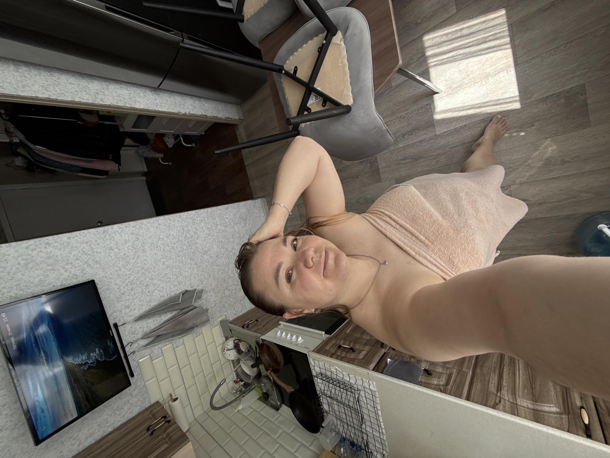 This is a high-angle, full-body, selfie-style photograph taken indoors, likely in a kitchen or living area. A woman, with wet hair and wrapped in a light pink towel, is the central subject. She is looking directly at the camera with her right hand resting on her head and her left arm bent. She is wearing a delicate silver necklace.
The environment appears to be a domestic setting with a kitchen counter to the left, featuring a sink, dishes, and cookware. A stovetop is visible behind the counter. A television is mounted on a wall above the kitchen area, displaying a scenic landscape. To the right of the woman, a dining table with two chairs is partially visible. One chair is overturned, with its legs in the air. The flooring is a light wood laminate. In the background, a doorway leads to another room where clothes are hanging on a rack, and a refrigerator is visible. The lighting suggests it is daytime, with sunlight casting striped shadows on the floor. The overall impression is a casual, perhaps post-shower, moment captured in a home.
The provided information about the location in the city of Stary Oskol, Russia, is external context and not discernible from the image itself. Therefore, this description is solely based on the visual evidence within the photograph.