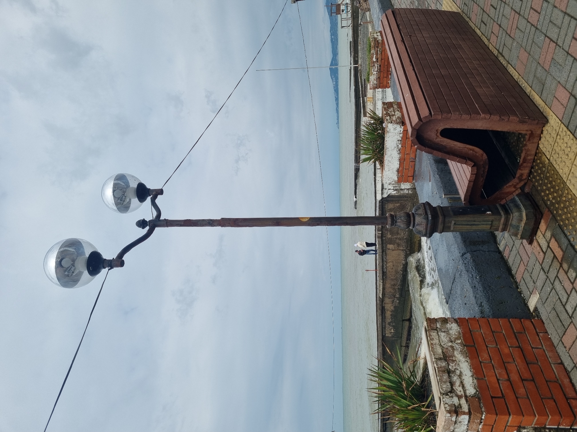 The image captures a seaside promenade in Sochi, Russia, on what appears to be an overcast or hazy day. In the foreground, a tall, antique-style streetlamp with a weathered, possibly rusted pole and two spherical glass globes stands prominently. Its base is ornate and dark.
To the right of the streetlamp, a modern, dark brown wooden bench with a distinctive wavy, ergonomic design provides seating along the walkway. The ground is paved with a mix of red and gray rectangular bricks in a patterned arrangement, alongside yellow tactile paving blocks closer to the bench. A low brick planter, partly whitewashed, holds spiky green plants, separating the paved area from a concrete barrier bordering the water.
Beyond the barrier, the sea stretches out under a light grey, cloudy sky. The water appears calm, with gentle waves breaking near the edge where the promenade meets the sea. In the middle distance, two small, indistinct figures, likely people, are visible walking along the concrete edge overlooking the water. The horizon is hazy, with faint outlines of distant land or mountains visible across the expansive sea. Several thin wires or cables crisscross high above the scene.
The overall atmosphere is quiet and serene, suggesting a peaceful moment on the coastal promenade. No discernible text is visible in the image.