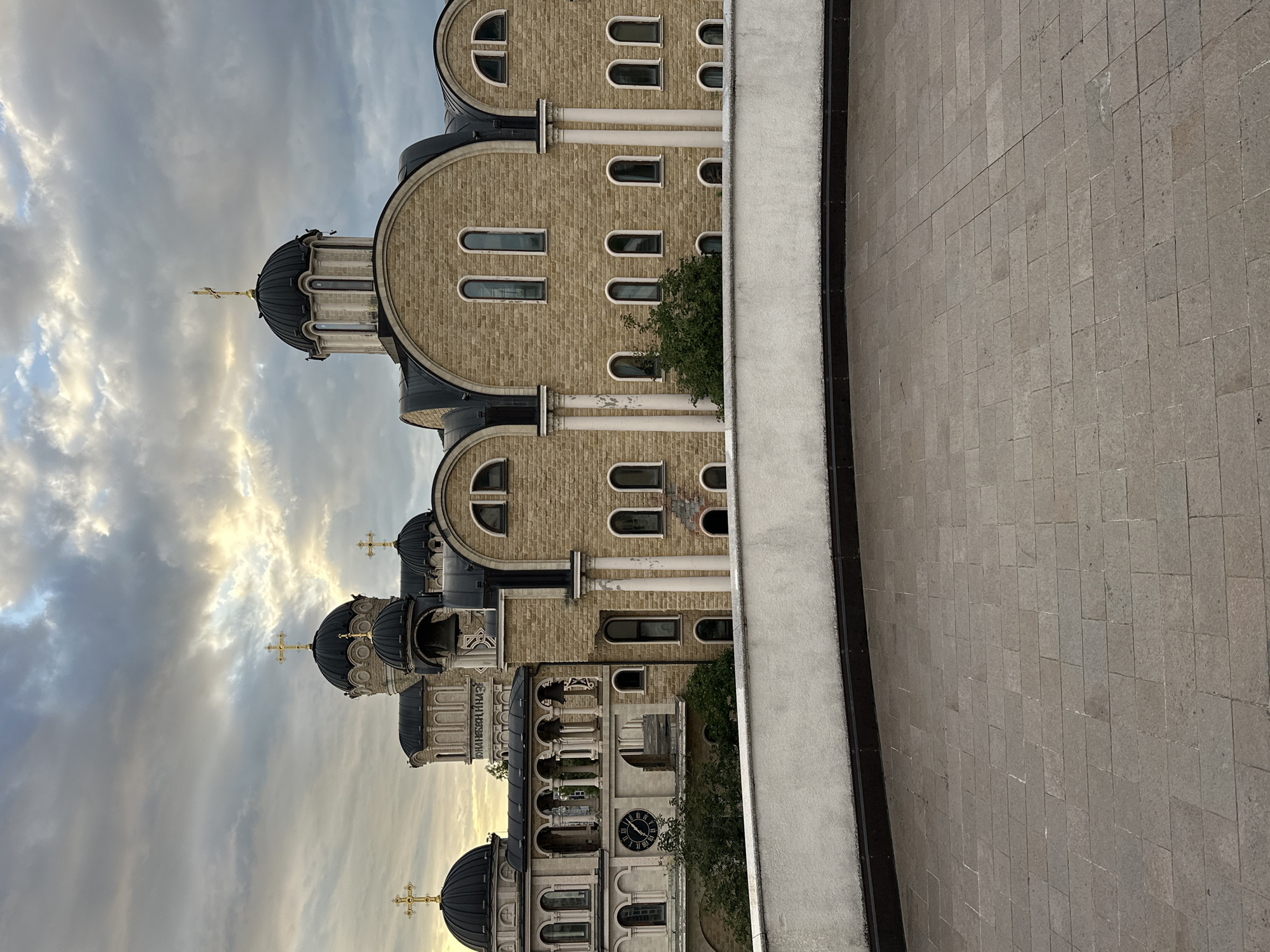 The image captures a view of the Church of the Transfiguration in Sirius, Russia, under a cloudy sky. The building features multiple domes topped with golden crosses and is constructed from light-colored stone. Arched windows and decorative columns are visible along the facade. In the foreground, there is a paved area and a low white wall. The overall scene suggests a late afternoon or early evening setting, as indicated by the lighting and cloud formations. There are no people visible in the image.