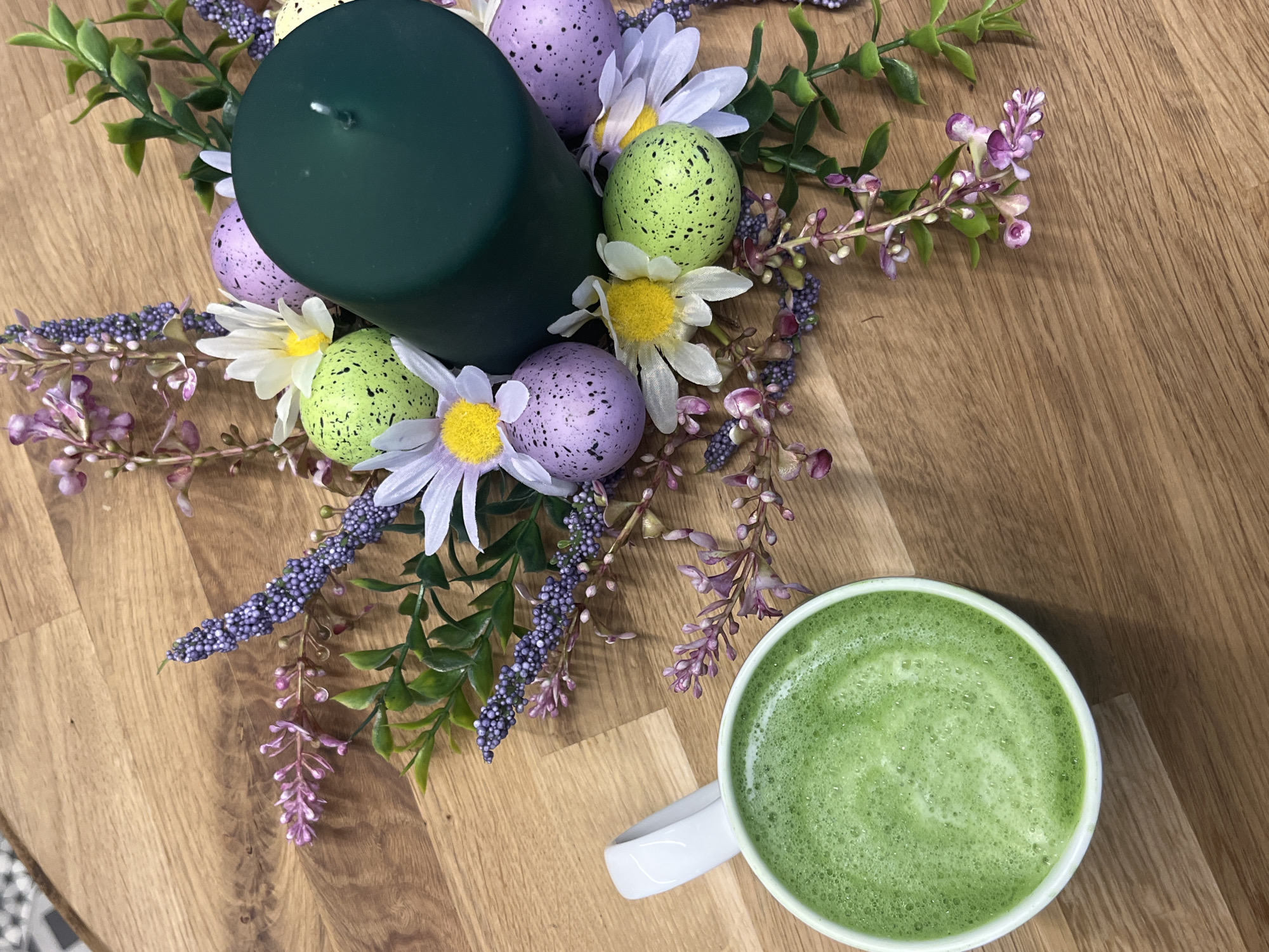 A high-angle shot depicts a table with a still life arrangement. The table has a light-brown, wood-grain surface. In the upper-left area, a round, dark-green candle is surrounded by a decorative arrangement of artificial elements. These include speckled Easter eggs in colors of green and lavender, white daisy-like flowers with yellow centers, green leaves, and sprigs of purple and lavender flowers. In the lower-right corner, a white ceramic mug is filled with a foamy, bright-green liquid, likely a beverage. The scene gives a domestic, possibly festive, ambiance. There is no information to determine the location of the image to be in Sofia, Bulgaria.