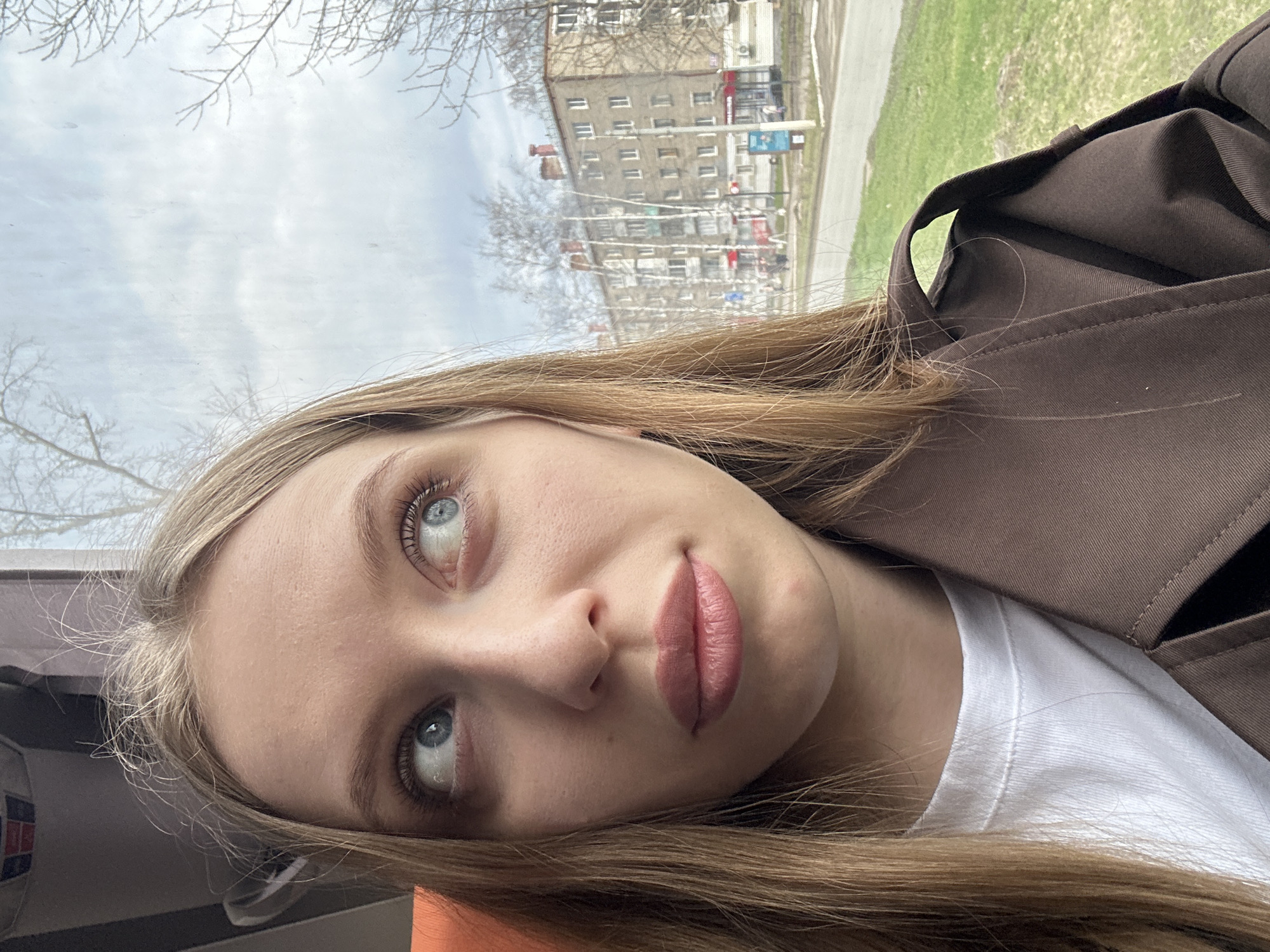 A close-up selfie of a young woman with light blonde hair and blue eyes. She is wearing a white t-shirt and a brown trench coat. The background shows a cityscape reflected in a window, including buildings and greenery. Based on the architectural style and other visual cues, the location is likely Chaikovsky, Russia.