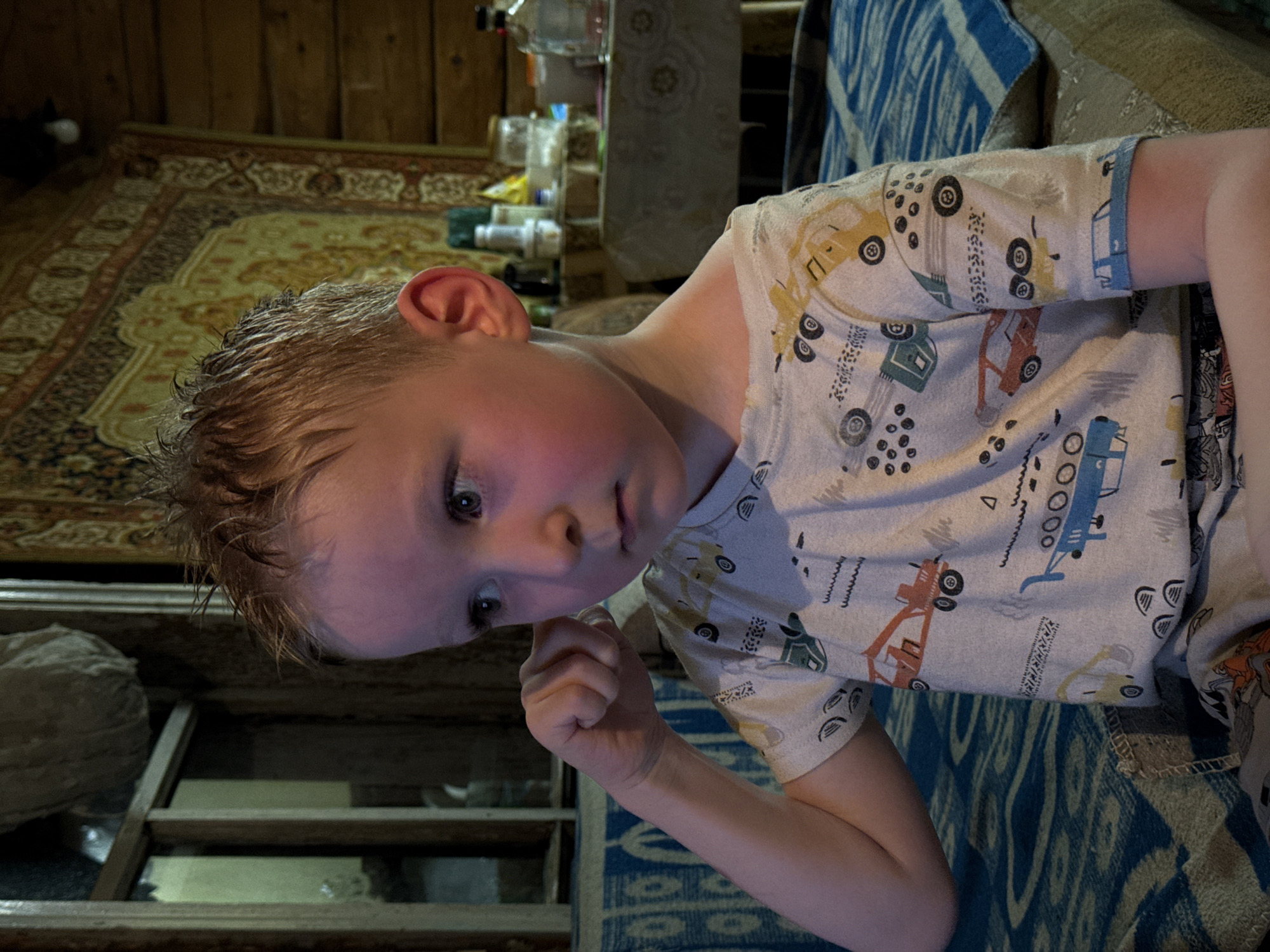 A young child with fair skin and wet blonde hair, wearing a white T-shirt patterned with vehicles, is lying down on a blue patterned blanket. The child is looking towards the camera with a slightly curious expression, and their left hand is raised near their face. The background consists of what appears to be a wooden interior, including shelves and a patterned rug. There are various items on a table in the background as well, including bottles and containers. While the location provided suggests the scene is in Nizhnyaya Chermoda, Russia, this is not verifiable from the image itself.