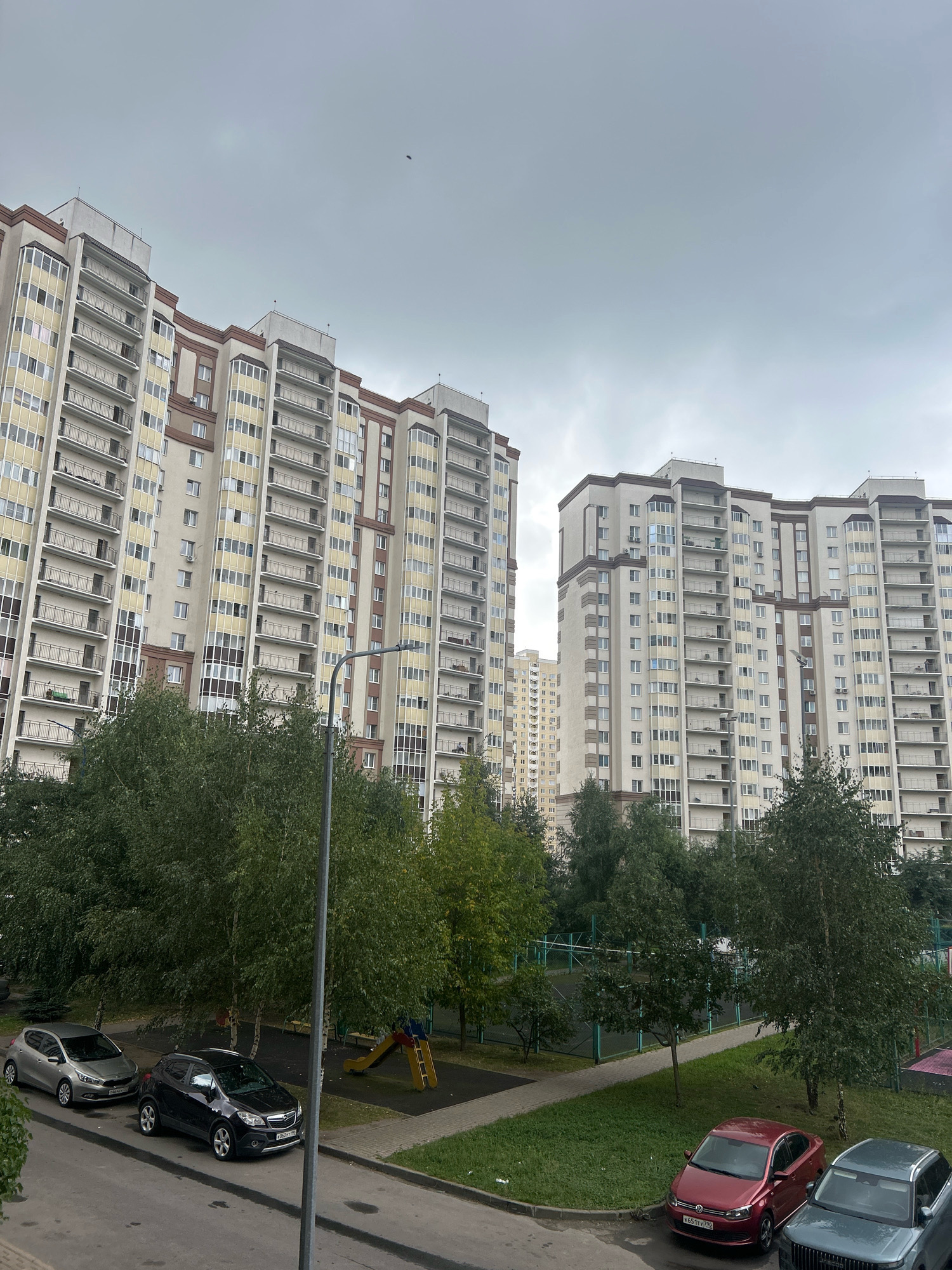 This image captures a residential complex in Domodedovo, Russia, under an overcast sky. Two multi-story apartment buildings dominate the scene, with their light-colored facades and numerous balconies. The buildings appear to be modern, with a mix of cream, beige, and brown architectural elements. In the foreground, a street is lined with parked cars, including a silver hatchback, a dark SUV, a red sedan, and a dark gray SUV. Adjacent to the street is a grassy area with trees, and a children's playground with a slide. Further back, a fenced sports court is visible. The overall impression is one of a typical urban residential environment.