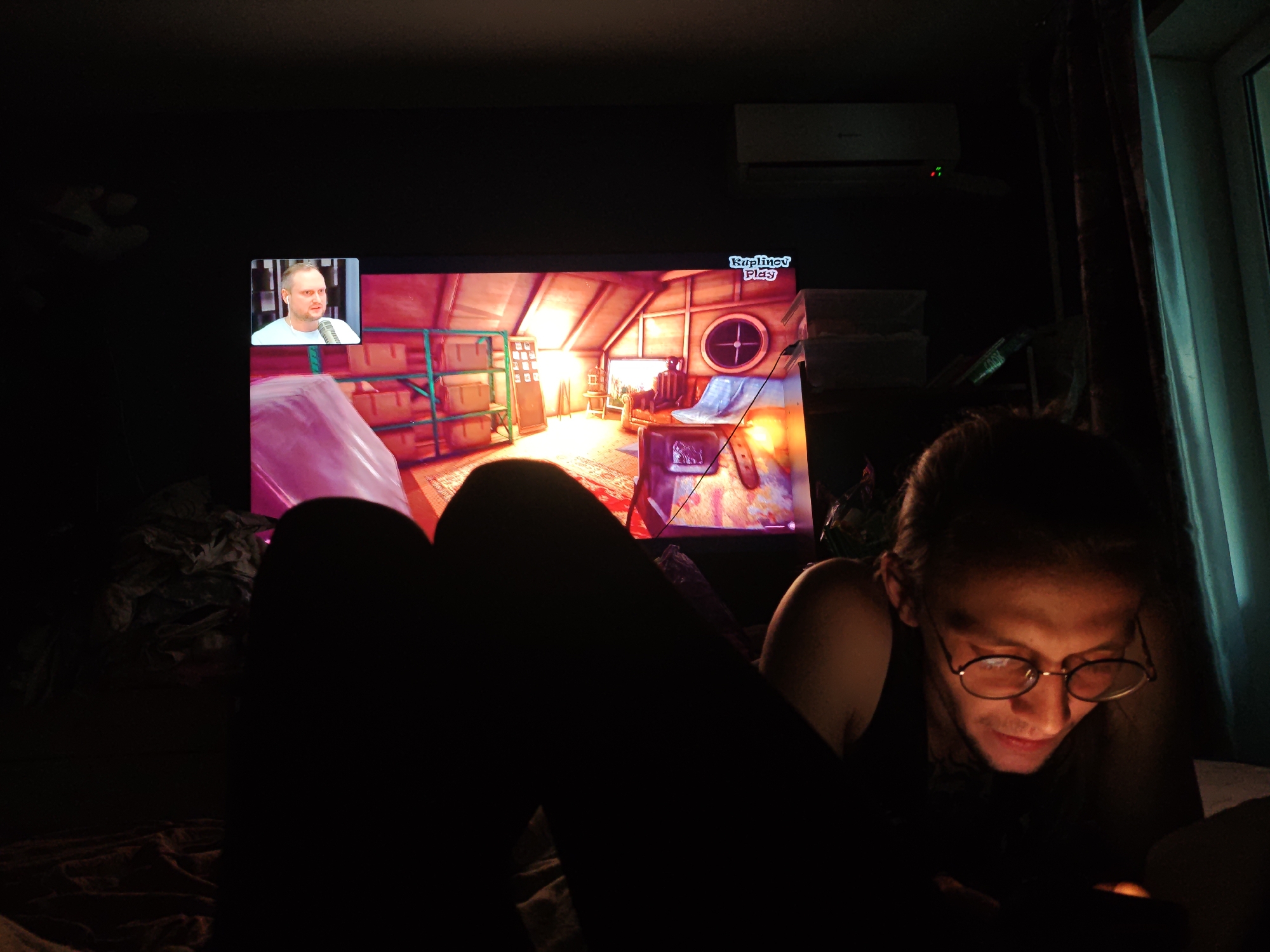 In a dimly lit room, an individual wearing glasses is looking down at a mobile device, their face illuminated by its screen. Behind them, a television displays a video game with a small inset window showing a man speaking. The room appears to be a living space with dark curtains and scattered objects. The specific location in Voronezh, Russia, cannot be determined from the image alone.