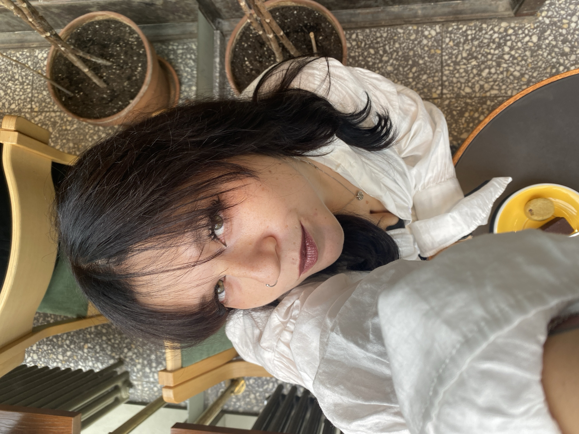 An overhead, close-up shot shows a woman with dark hair and light green eyes. She is wearing a white collared shirt with puffy sleeves and a silver necklace. Her lips are painted with a dark plum lipstick, and she has a silver nose ring. The woman is looking up at the camera with a soft expression. She is seated at a dark brown table with a yellow plate holding two small cookies. The background shows a floor with a speckled terrazzo pattern and two terra cotta pots with sparse branches. The scene appears to be an outdoor seating area of a cafe. The style of the cafe, with its terrazzo flooring and outdoor seating, is common in Saint Petersburg, Russia.