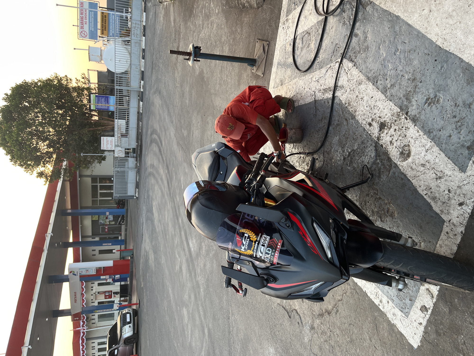 A person in an orange uniform and cap is crouching beside a black motorcycle at a Pertamina gas station in Pecangaan, Indonesia. The person appears to be cleaning or working on the motorcycle. The motorcycle is parked on a concrete surface marked with white lines. In the background, there is a gas station canopy with red and white trim, fuel pumps, and a store. A silver SUV is parked nearby. A large tree and signage are visible behind a fence. The sky is a light orange, suggesting it is either sunrise or sunset.